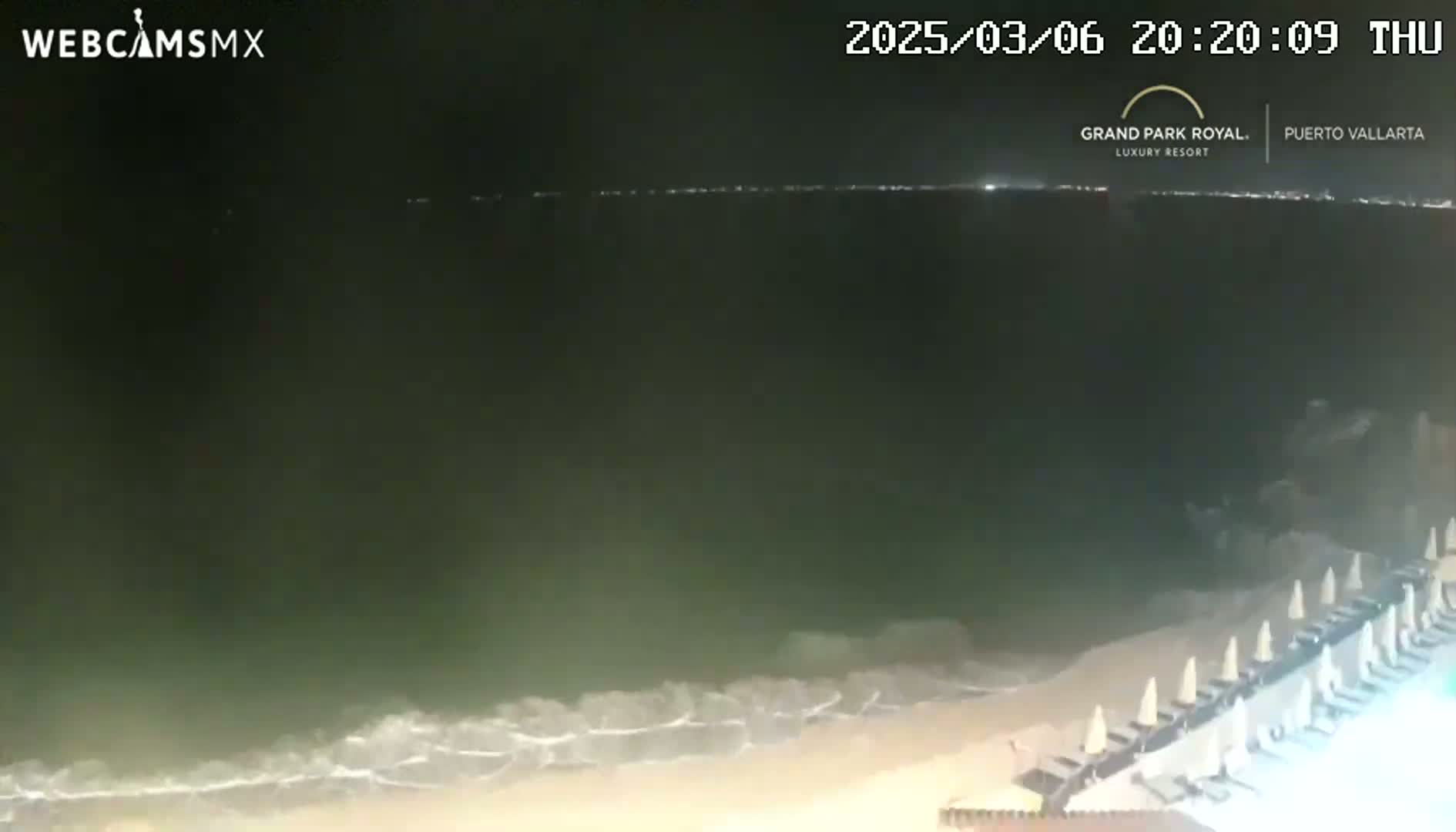 A nighttime beach scene shows gently lapping waves on a sandy shore lined with closed beach umbrellas.