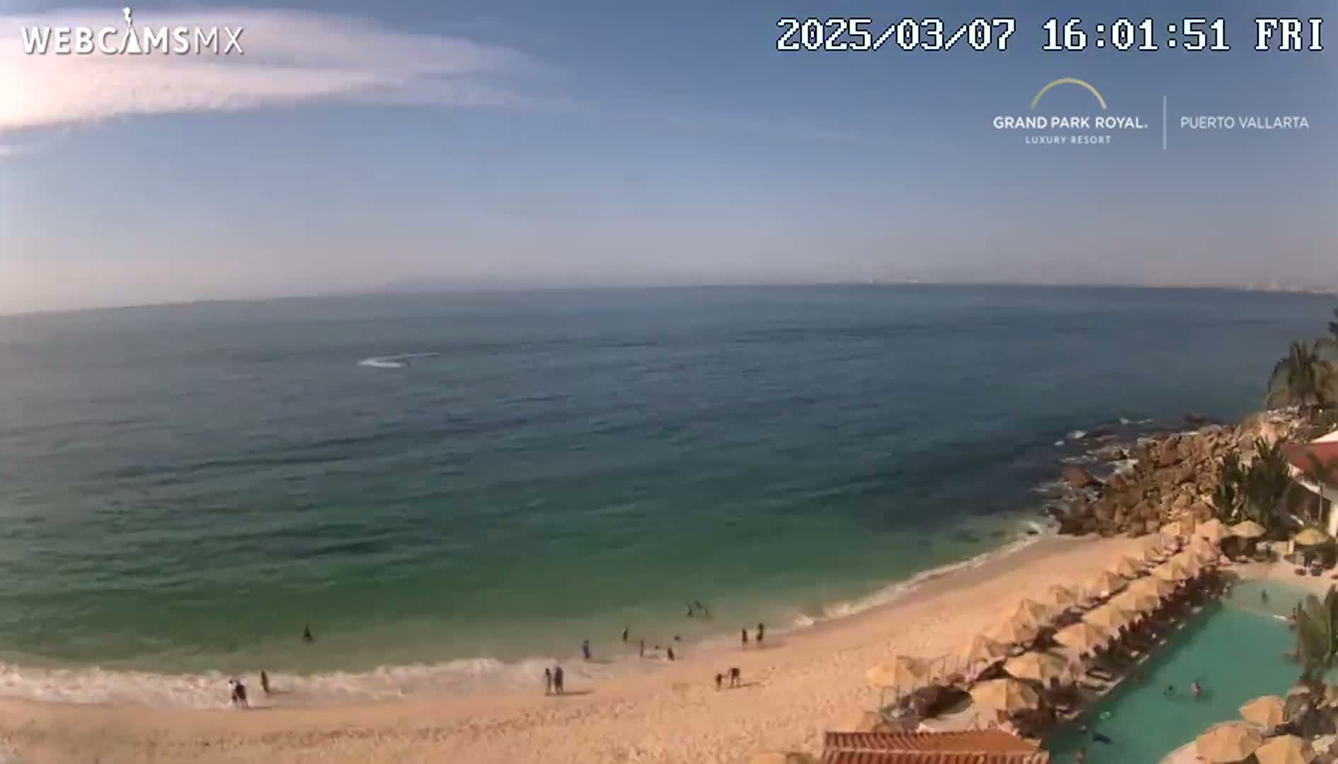 A mostly sunny day at a beach with calm, teal water, a sandy shore, and a resort with a pool and umbrellas.