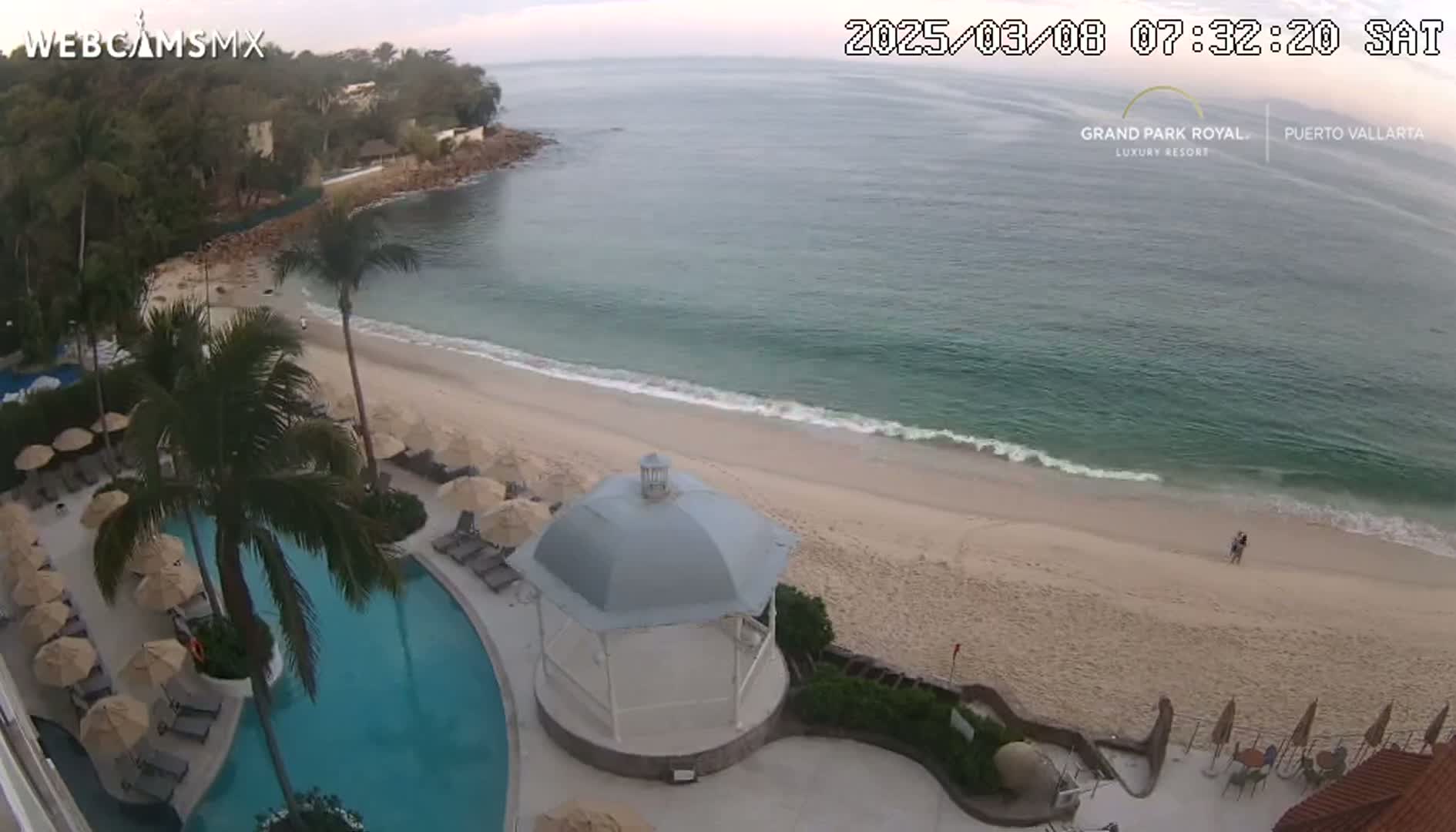 An aerial view shows a calm ocean lapping a sandy beach next to a resort with a swimming pool and gazebo, under an overcast sky.