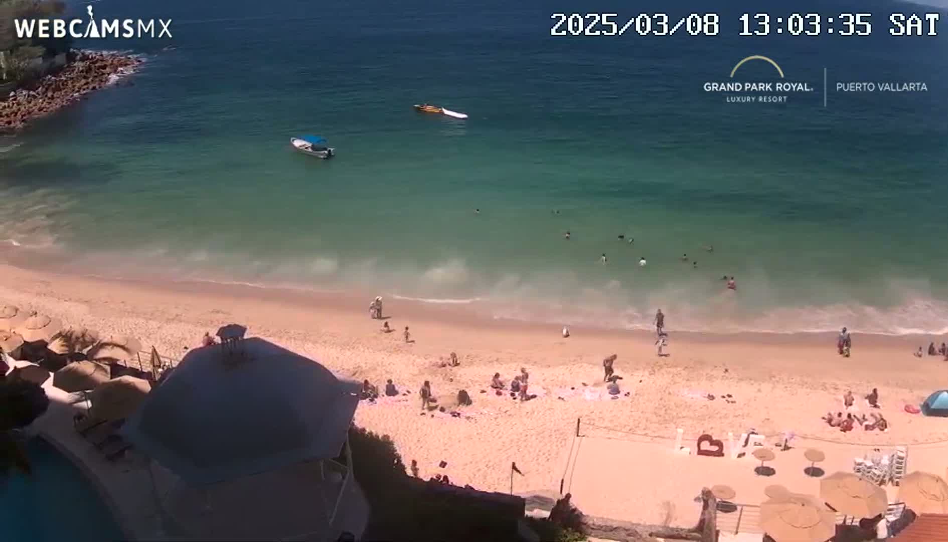 A sandy beach with people swimming and sunbathing, two boats in the turquoise water, and several umbrellas on the sand under a sunny sky.