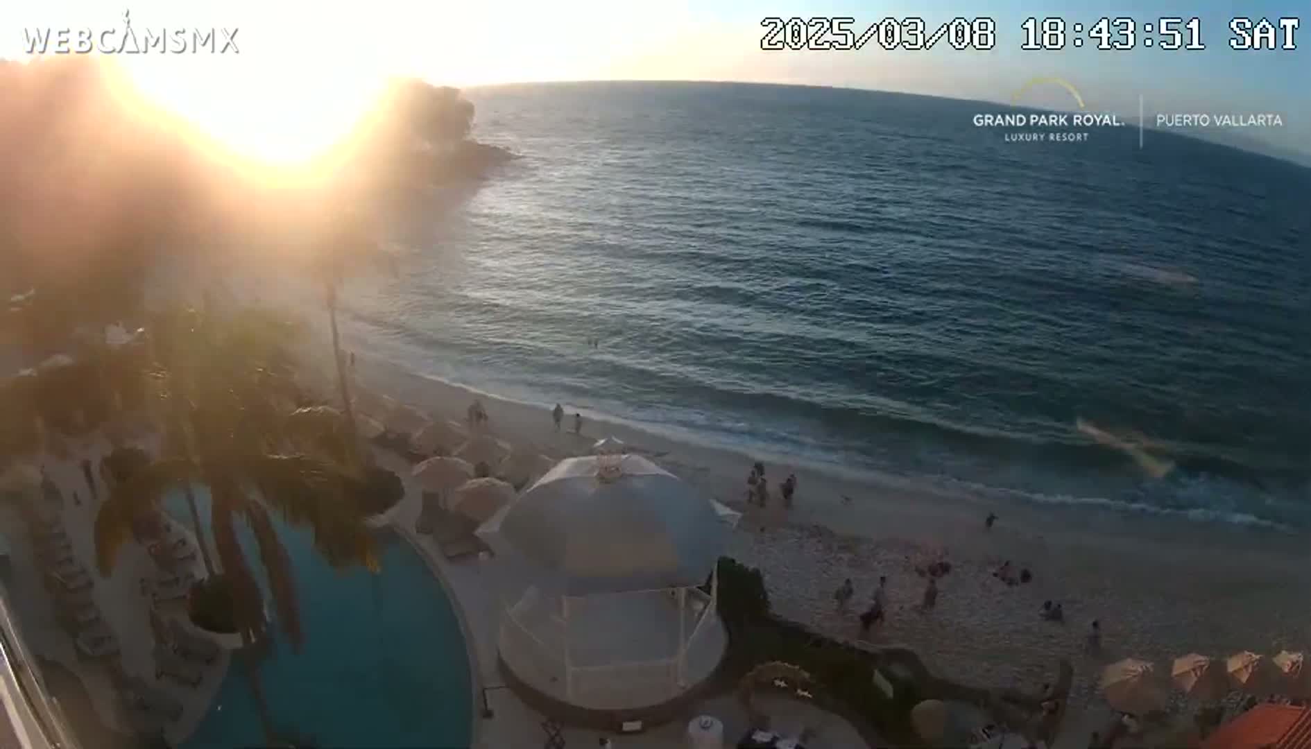 A beach scene at sunset shows a sandy shoreline with people relaxing, an ocean with gentle waves, a resort pool, and palm trees near a building.