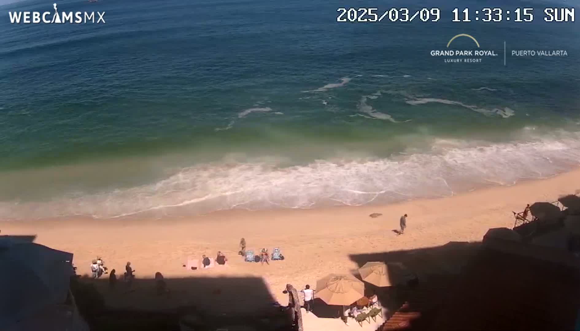 A sandy beach with a few people and beach umbrellas is seen from above, next to a calm ocean on a sunny day.
