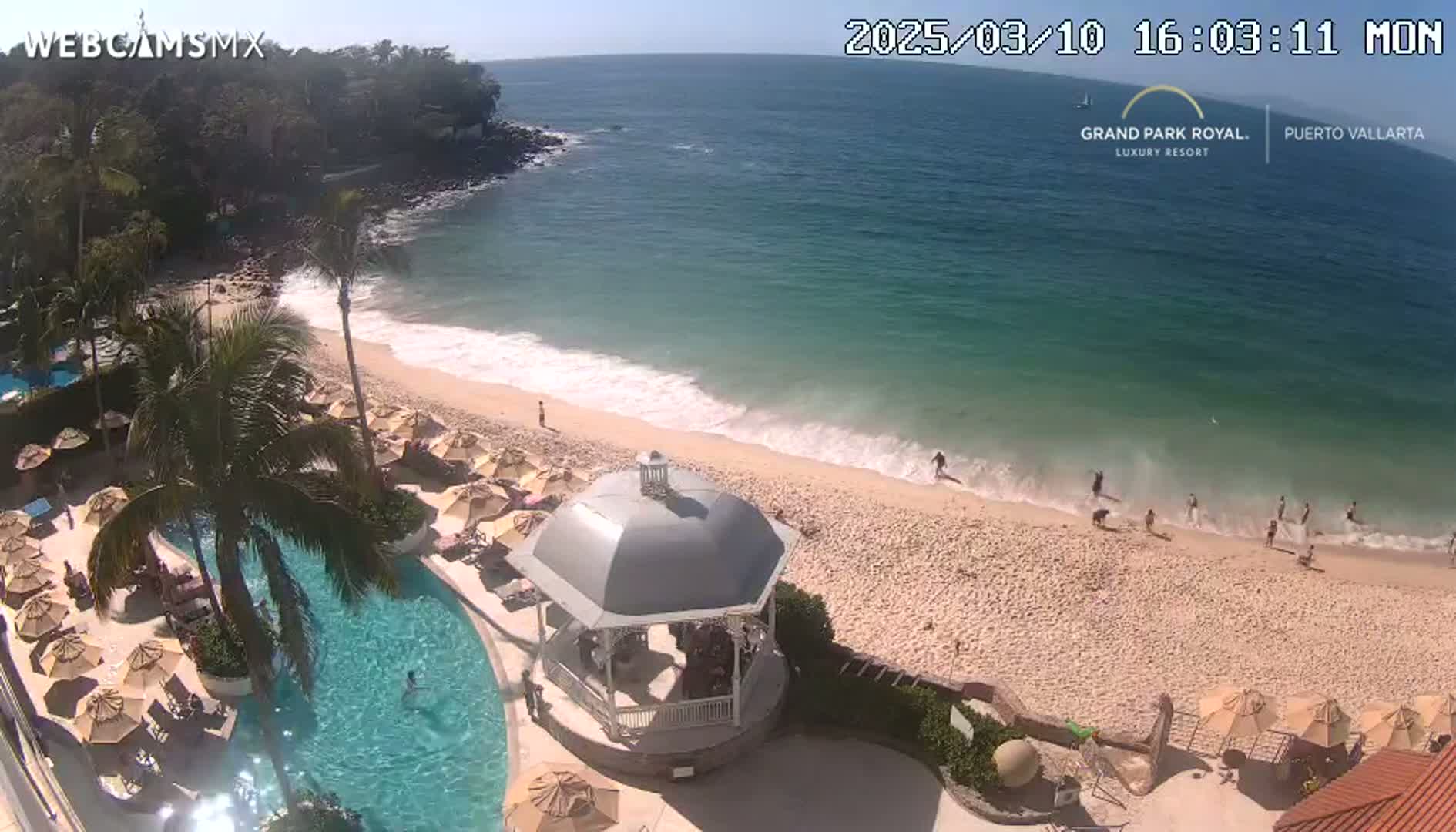 An aerial view shows a sandy beach with gentle waves lapping the shore,  people enjoying the beach and a resort pool area with a gazebo under a sunny sky.
