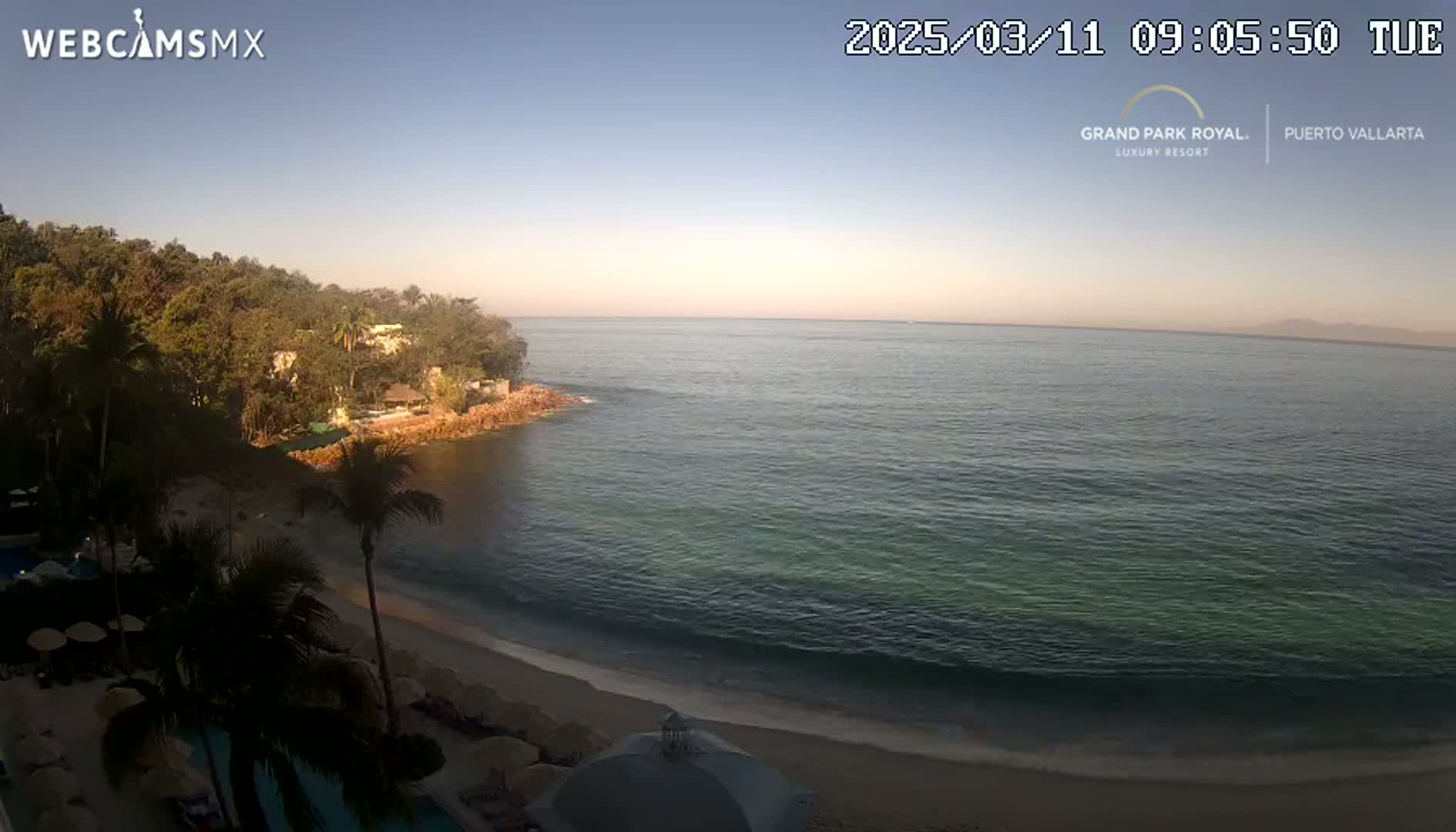 A tranquil beach scene at sunrise shows a calm ocean lapping a sandy shore, backed by lush vegetation and a resort.