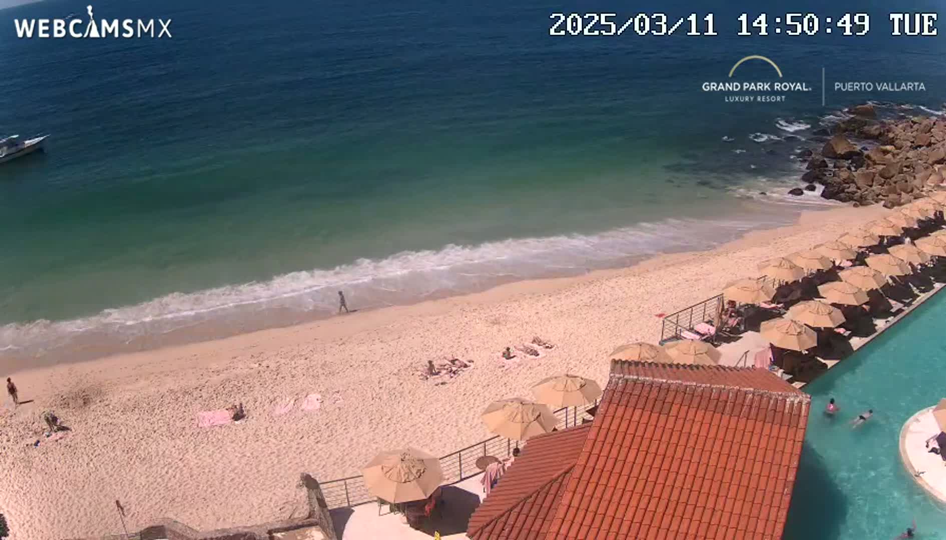 An aerial view shows a sandy beach with a few people on it, gentle waves lapping the shore, and a resort with a swimming pool and numerous beach umbrellas.