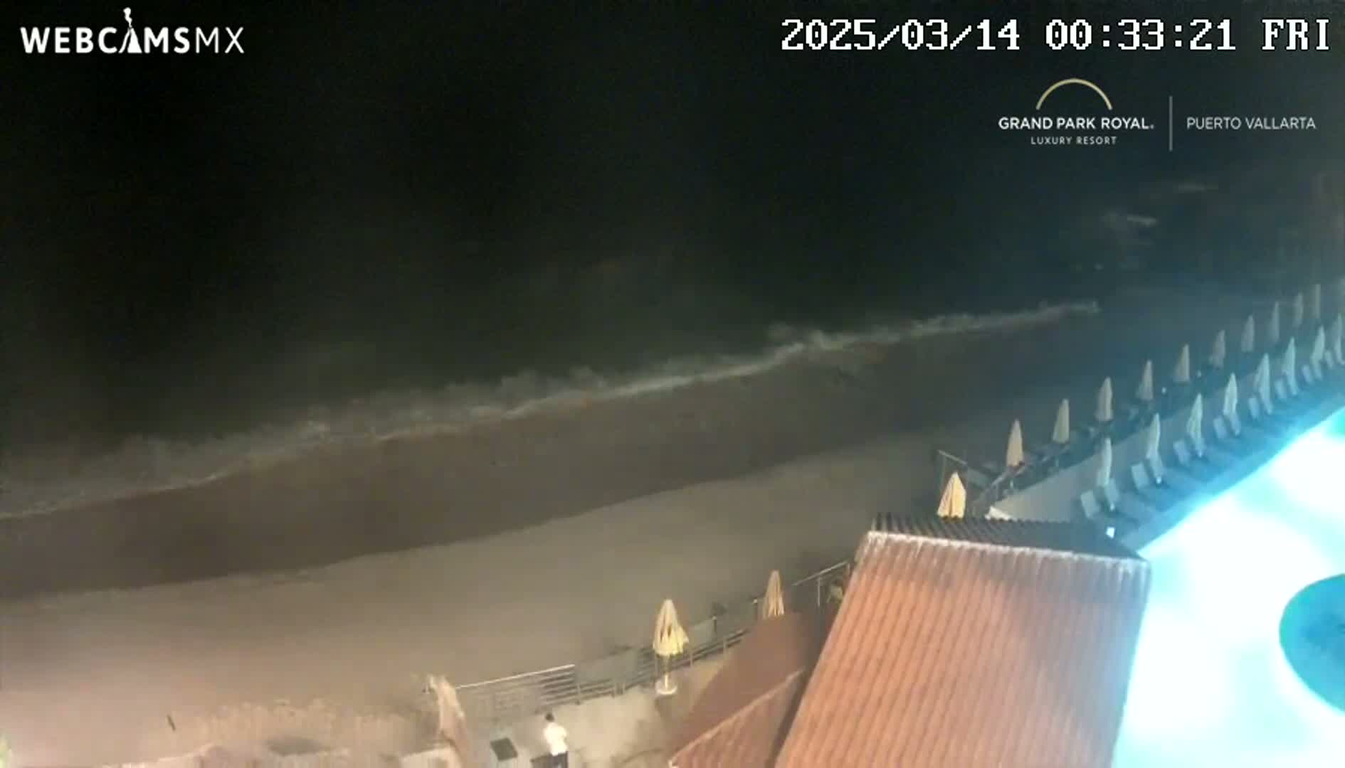 A nighttime beach scene shows dark waves lapping a sandy shore, with beach chairs and umbrellas lined up along a resort building illuminated by a bright blue pool light.