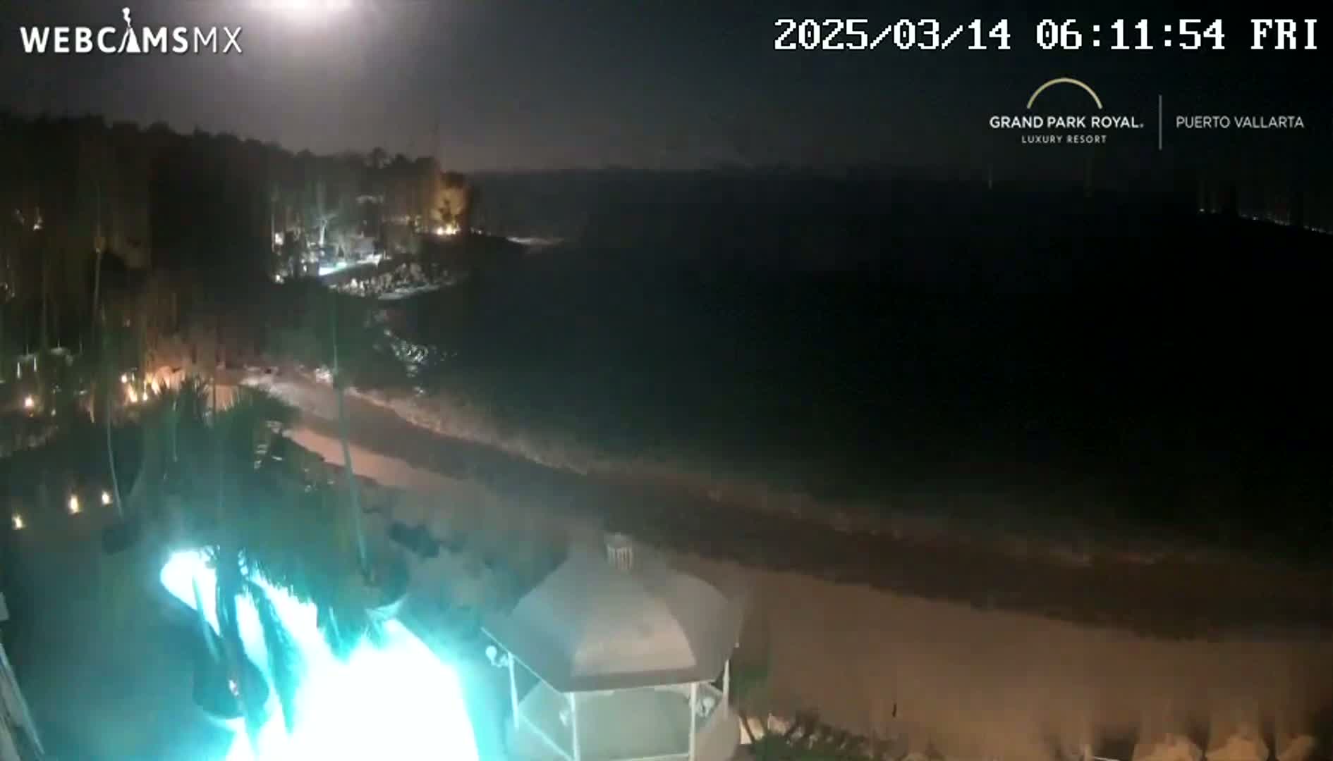 A nighttime view shows a resort pool lit in bright teal, a beach, and dark ocean under a clear, dark sky.