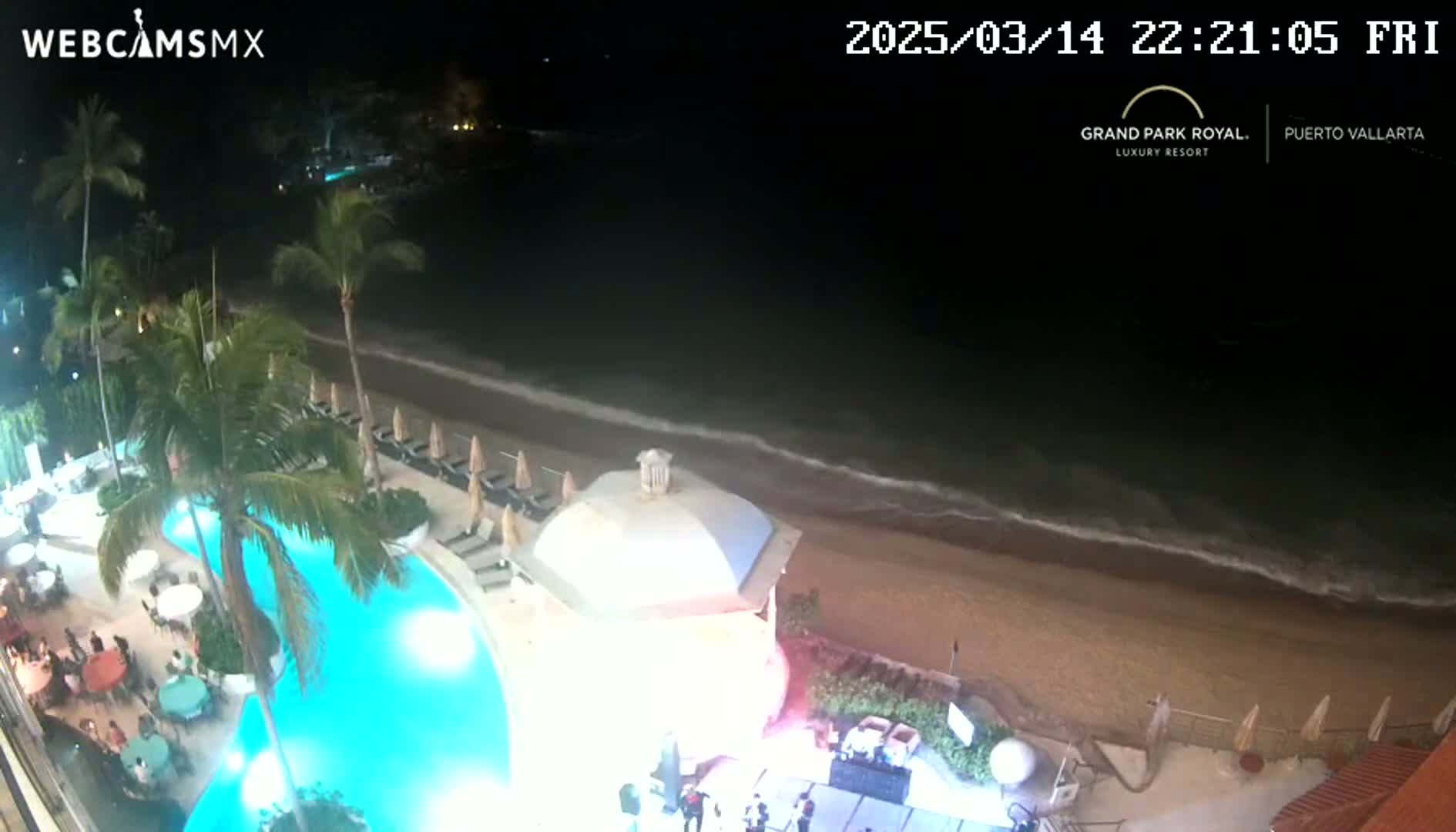 A nighttime aerial view shows a resort pool, beach, and ocean with gentle waves.