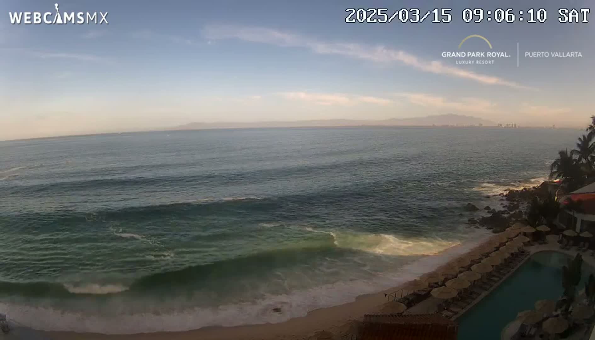 An ocean beach with moderate waves under a mostly clear sky, viewed from above a resort pool area.