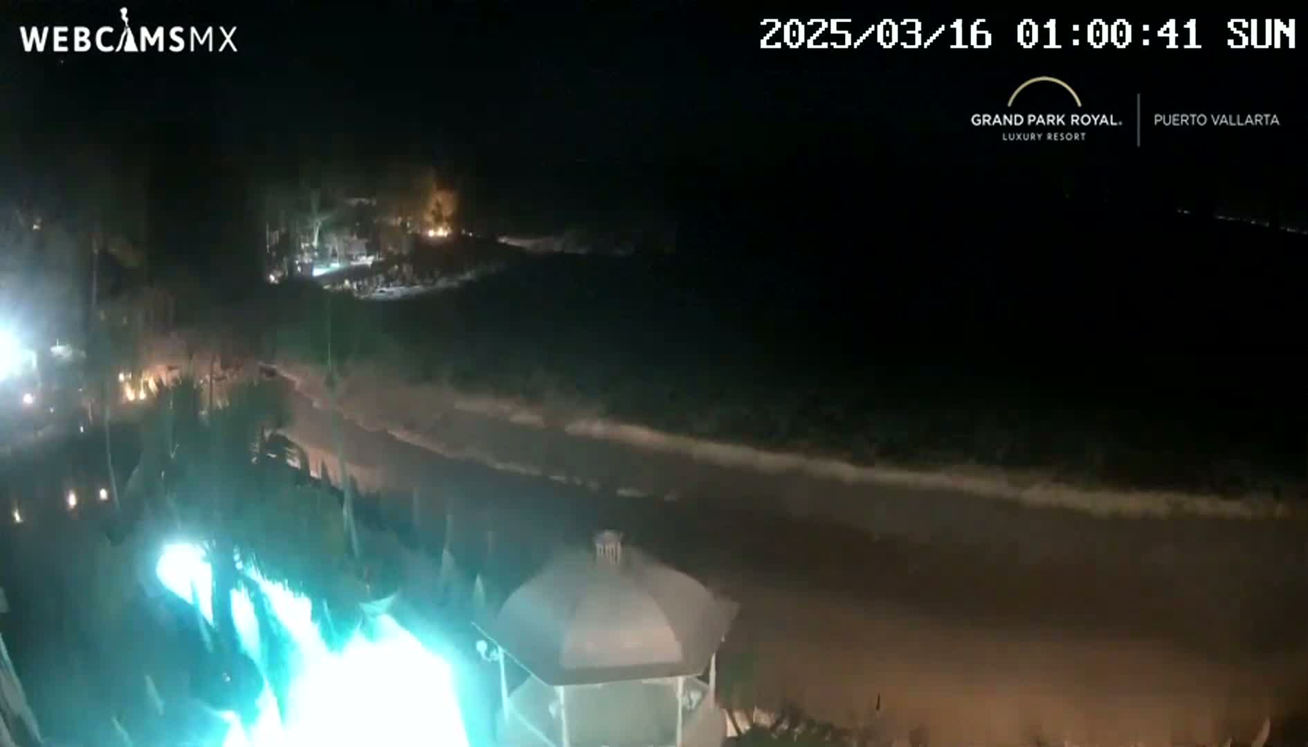 A nighttime beach scene shows a dark ocean with small waves lapping the shore near a gazebo illuminated by bright turquoise lights, with vegetation and some lights visible further inland.