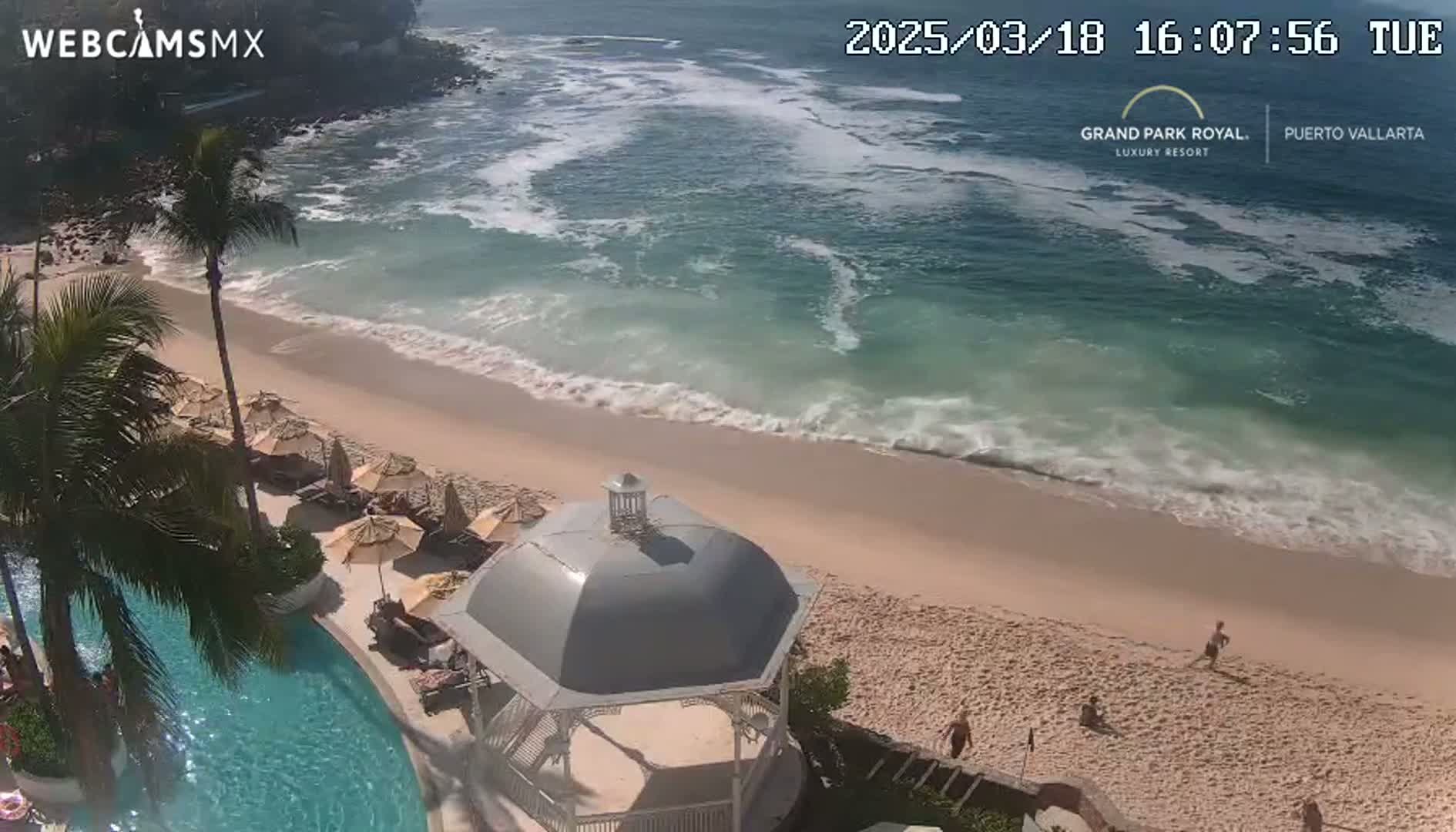 An aerial view shows a sandy beach with gentle waves lapping the shore, a resort pool, and a gazebo, under sunny skies.