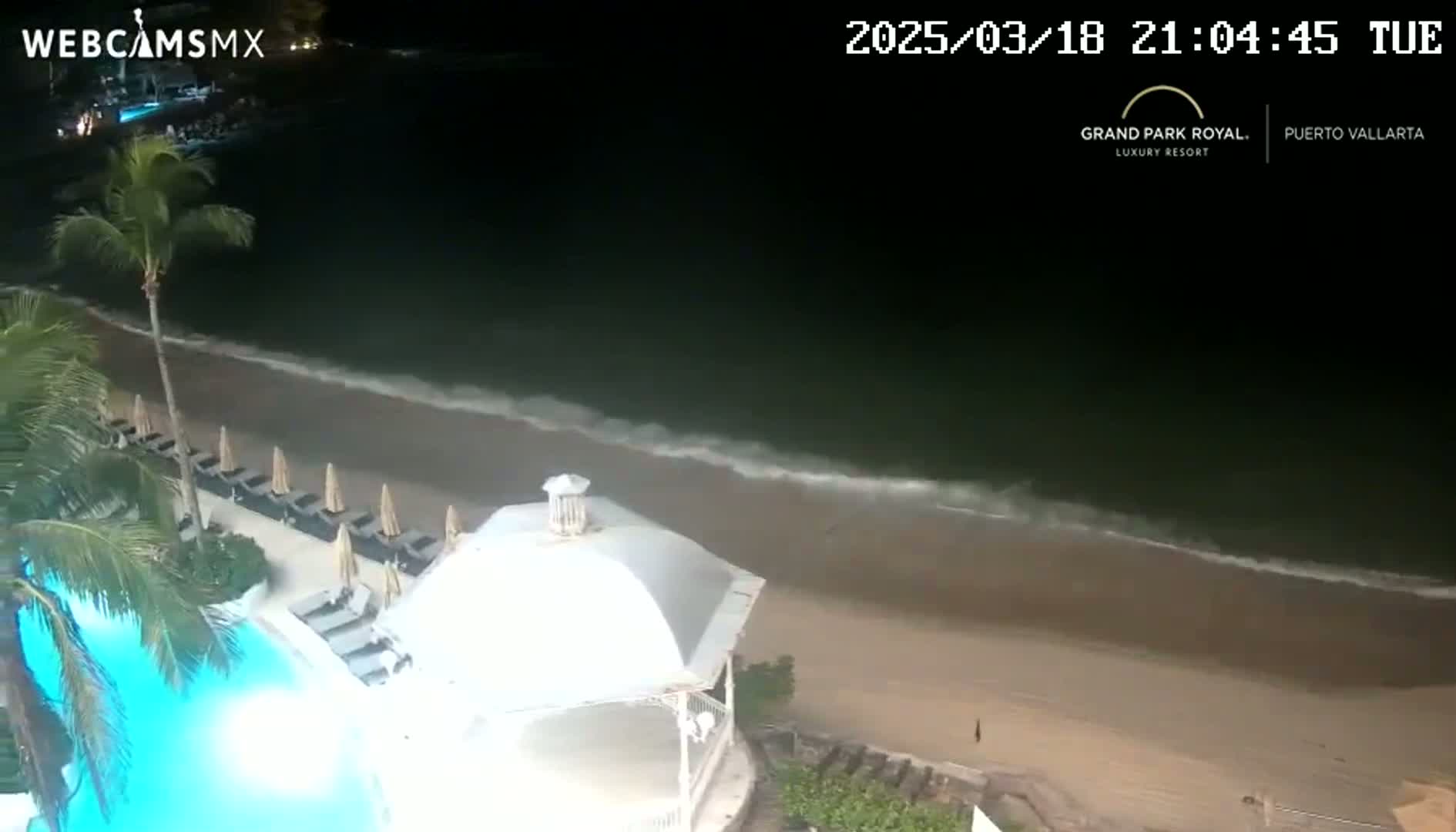 A nighttime, overhead view shows a resort pool and beach with gentle waves lapping the shore under a dark sky.
