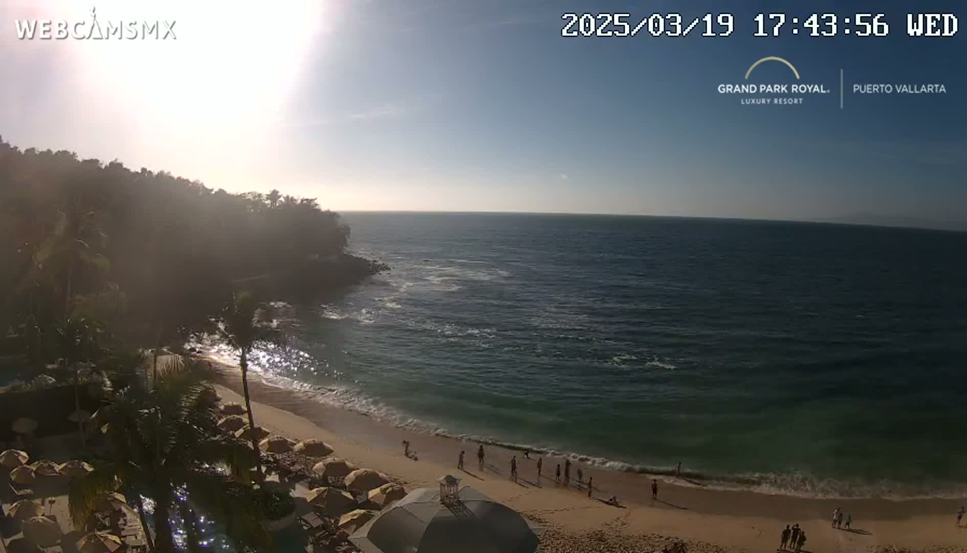 A sunny beach scene shows a sandy shore with gentle waves lapping at the edge,  people relaxing on the beach, and a resort with numerous umbrellas and palm trees near a calm ocean under a mostly clear sky.