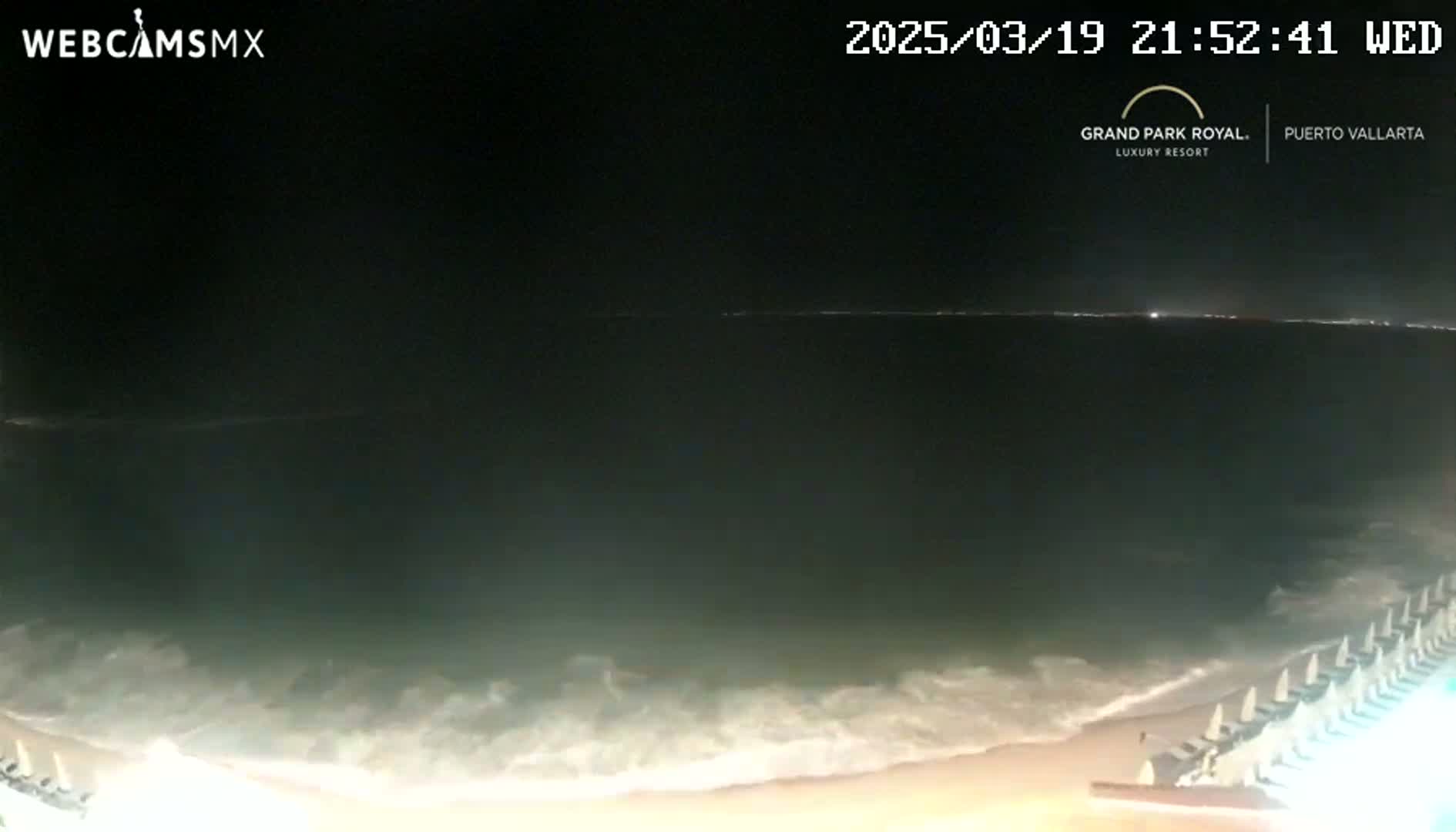 A nighttime beach scene shows dark ocean waves lapping a sandy shore with rows of beach chairs under a clear, dark sky.