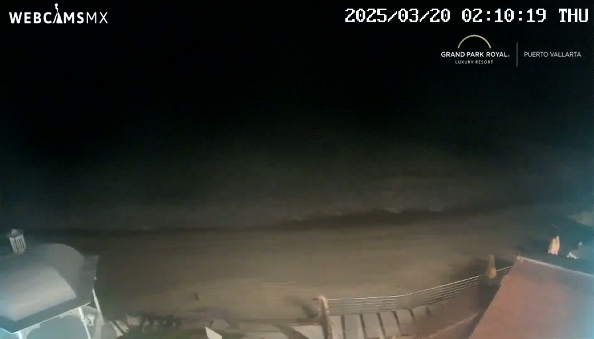 A dark, nighttime beach scene shows a mostly empty sandy shore with small waves lapping at the edge, and a portion of a building is visible in the lower right and lower left corners.