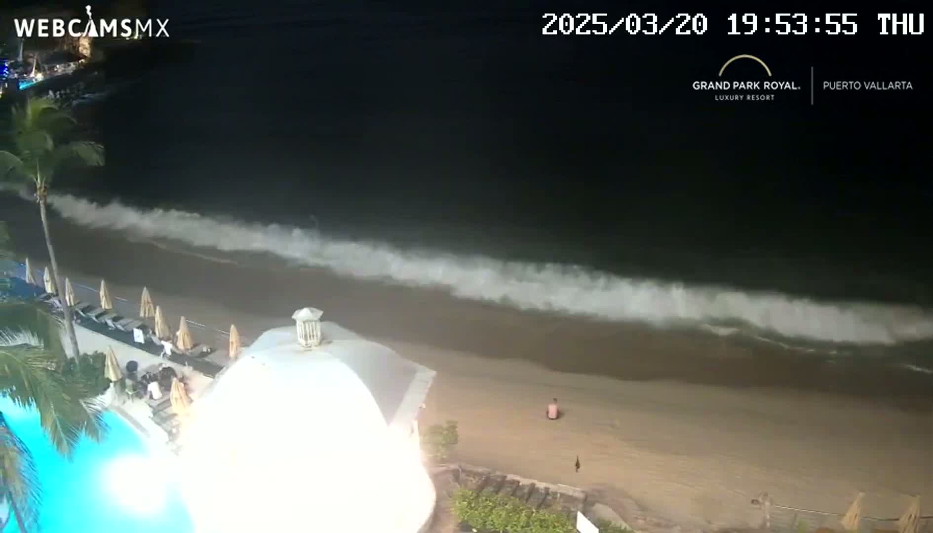 At night, moderate waves crash on a sandy beach next to a resort pool area with people present.