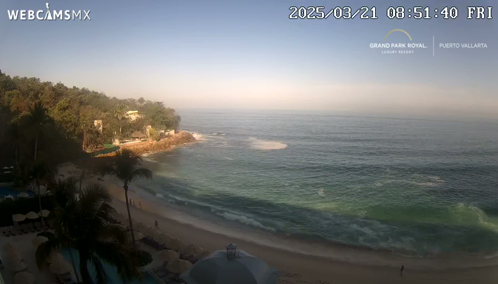 A beach with calm waves laps a sandy shore lined with palm trees and resort buildings under a clear, bright sky.