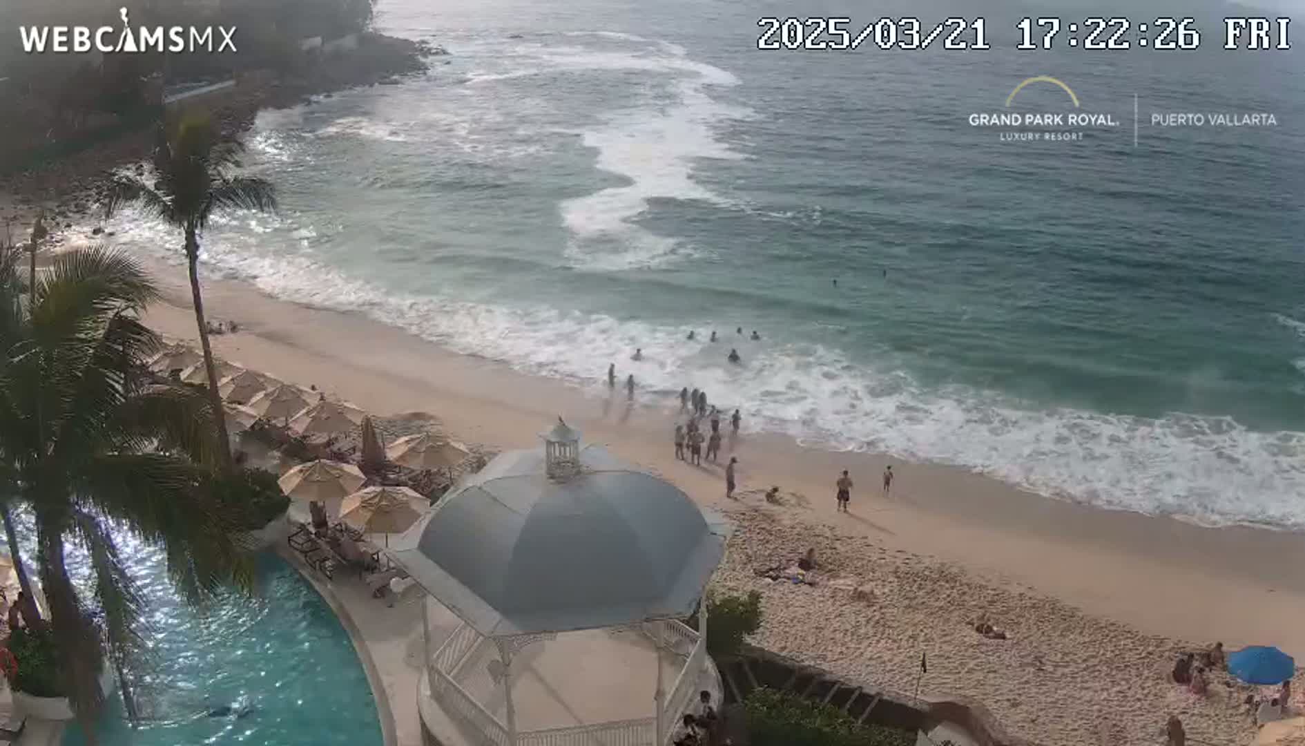 An aerial view shows a sandy beach with people swimming and relaxing near the shore,  under partly sunny skies, next to a resort with a pool and gazebo.