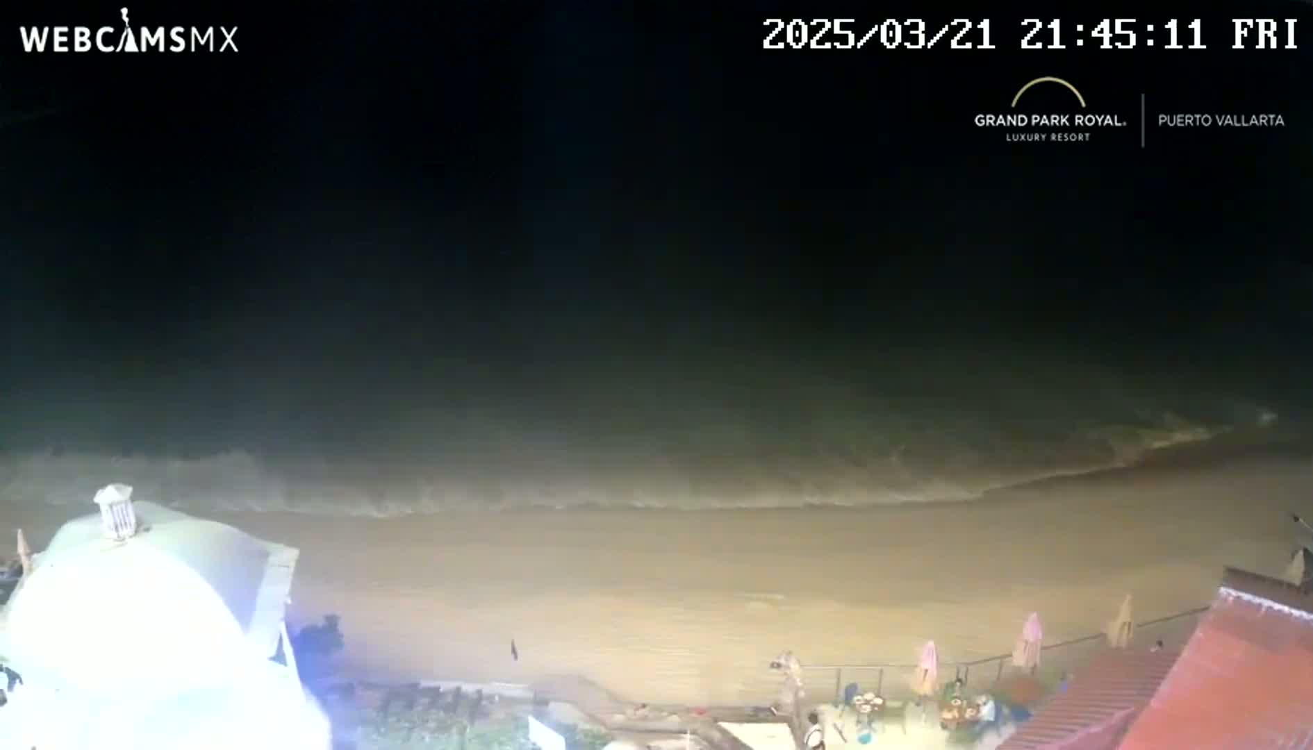 A nighttime beach scene shows gentle waves lapping at a sandy shore, with a few people visible near some beachside structures.