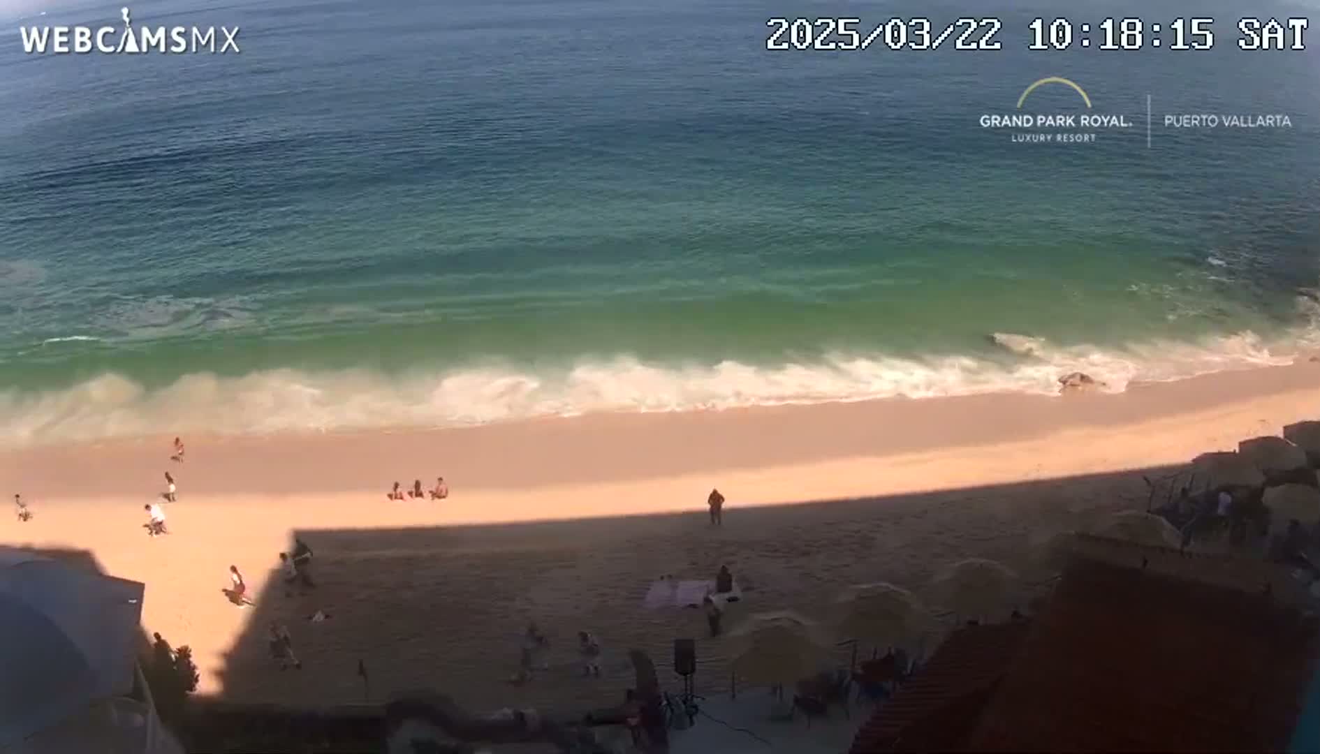 A beach with light-colored sand and turquoise water with gentle waves is partially shaded by a building, with several people scattered on the sand.