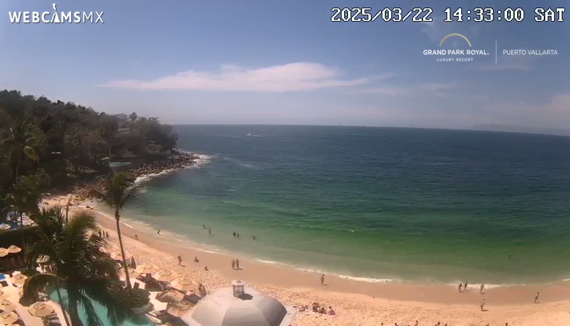A beach with clear, teal water and a sandy shore is viewed from above on a sunny day, showing people swimming and relaxing.