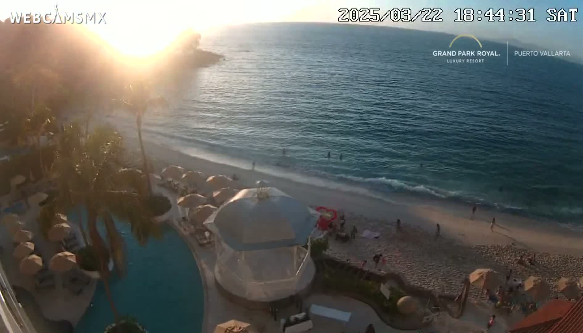 An aerial view shows a beach with calm waves at sunset, a resort pool, and palm trees, all under a clear sky.