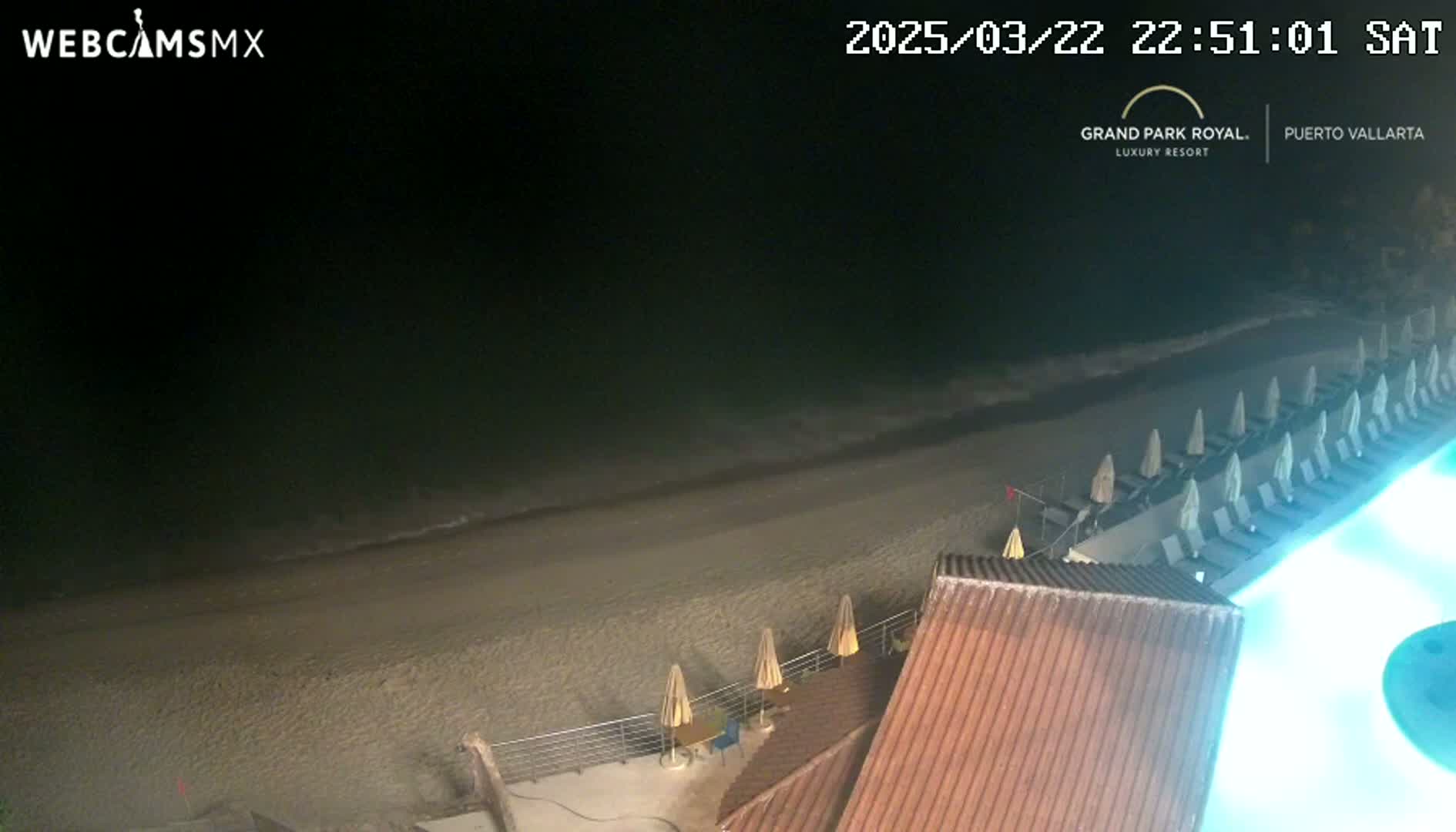 A nighttime beach scene shows a sandy shoreline with gentle waves, closed beach umbrellas and lounge chairs, and a portion of a building with a tiled roof next to a lit pool.