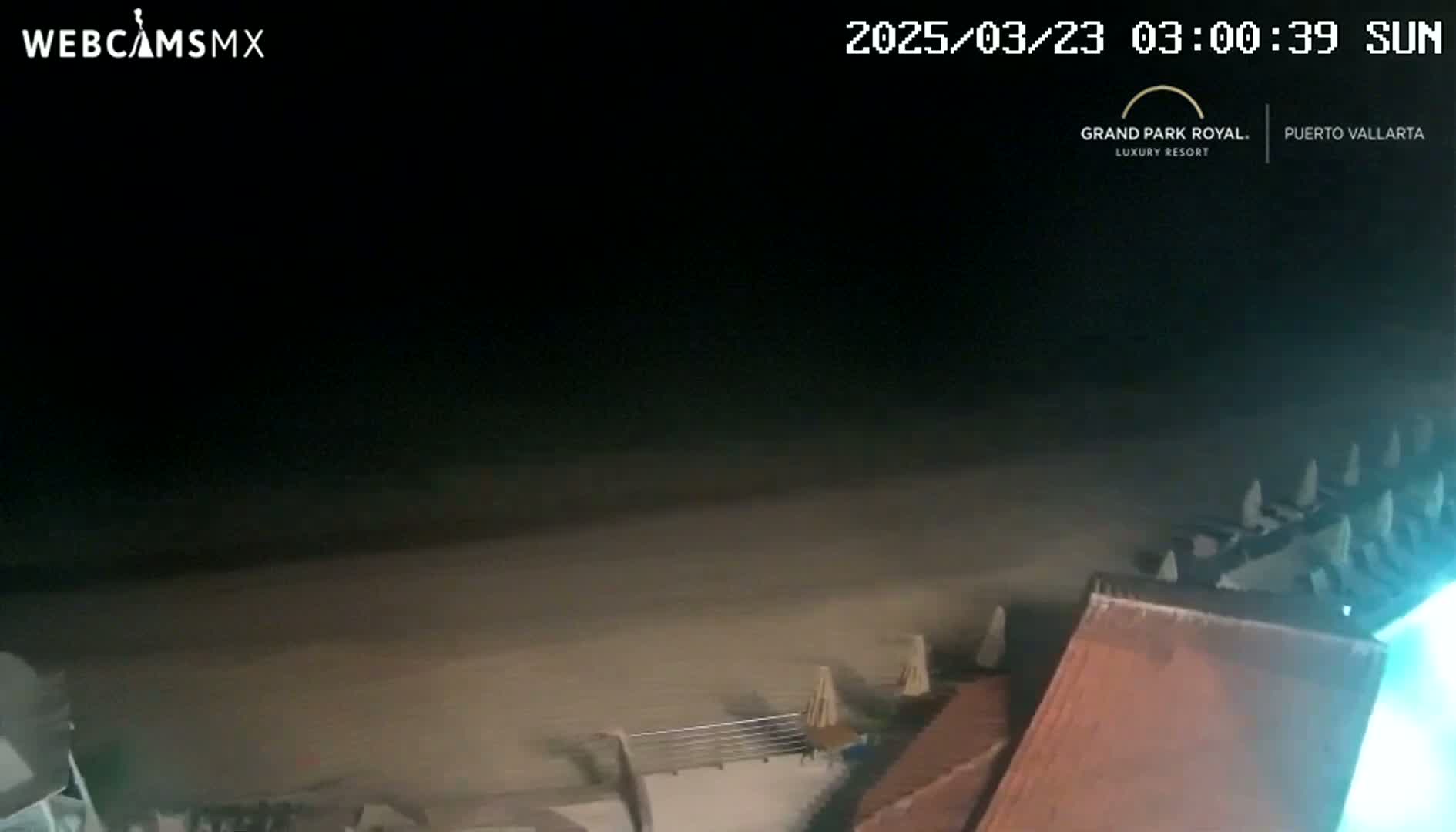 A dark nighttime beach scene shows a sandy shoreline with part of a building and some beach umbrellas visible in the lower right corner.