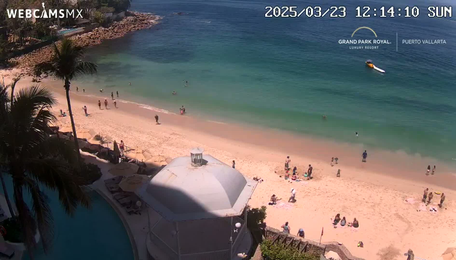 A sandy beach with many people swimming and sunbathing, situated next to a calm, teal ocean on a sunny day.