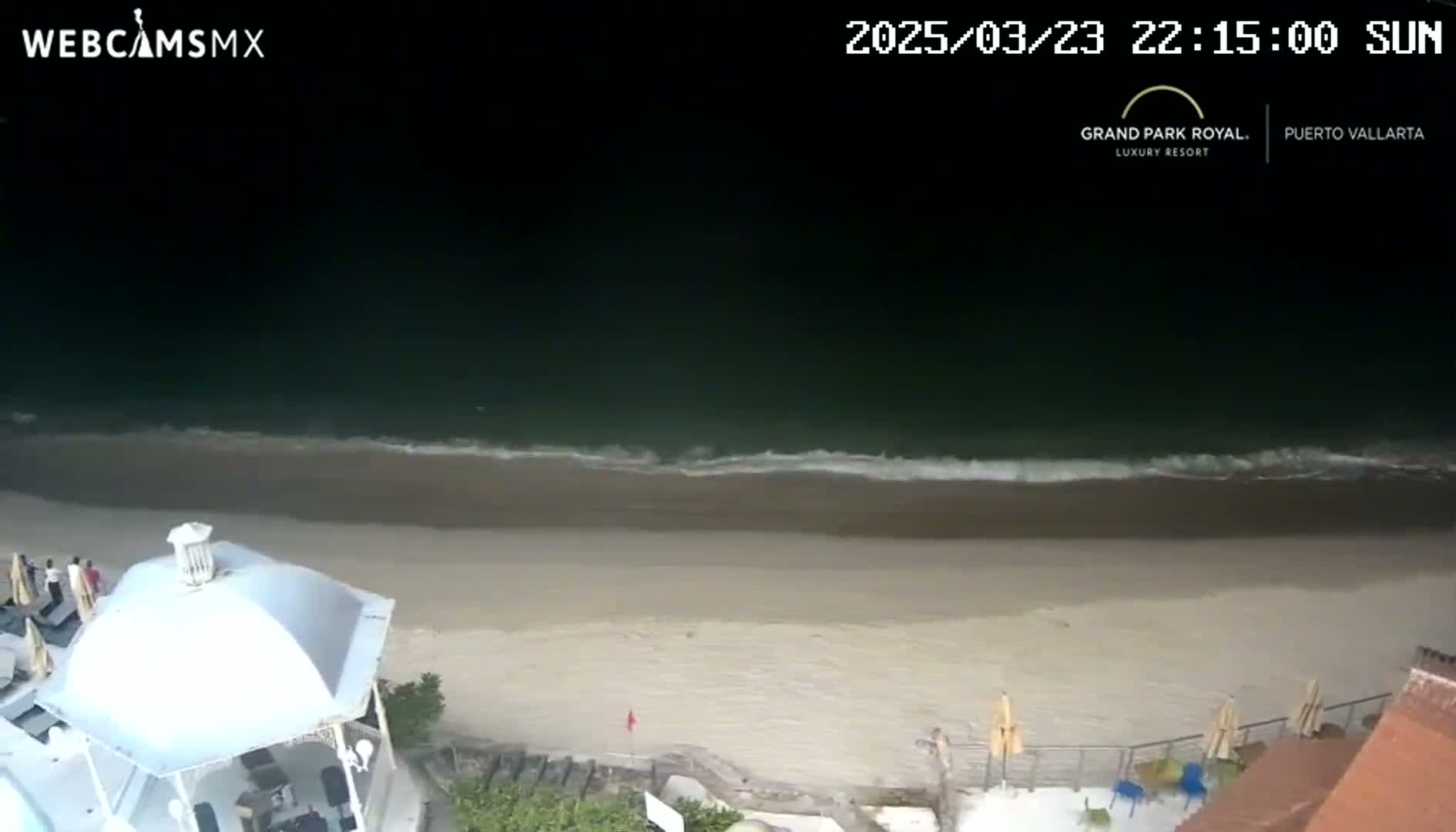 A nighttime beach scene shows a dark ocean with small waves lapping onto a light-colored sandy shore, partially visible from an elevated vantage point near a white gazebo.