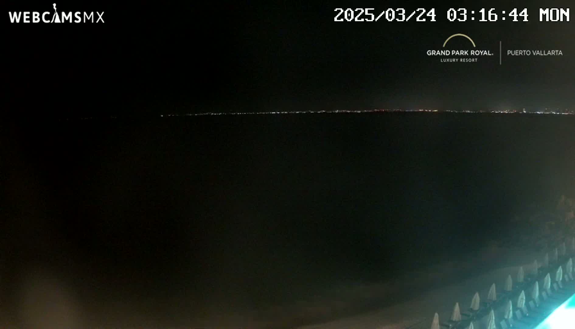 A nighttime view of a dark ocean with a distant city skyline illuminated, and beach chairs lined up on the sand in the foreground.