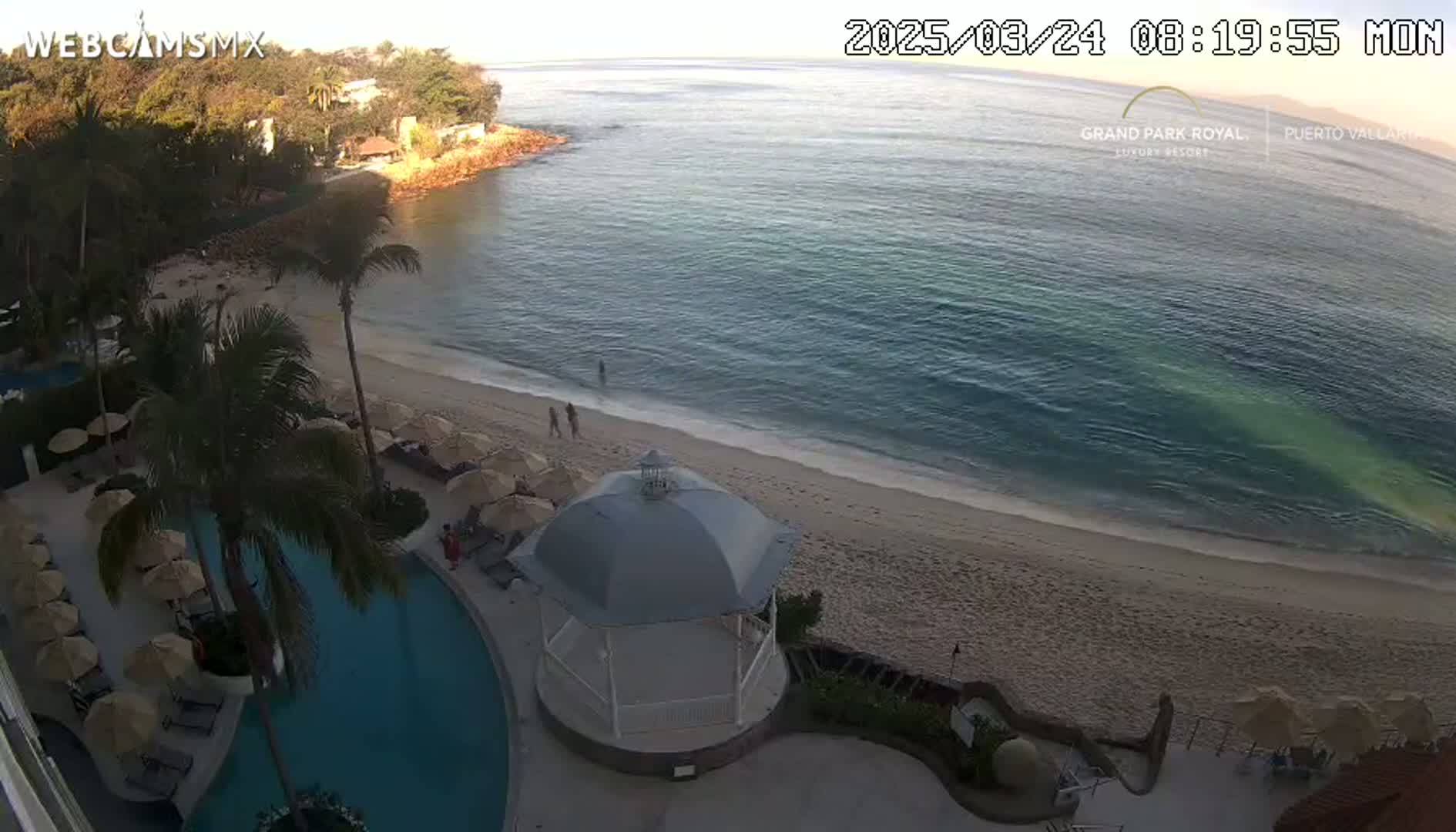 An aerial view shows a sandy beach meeting a calm, teal ocean under a clear sky, with a resort pool and gazebo visible near the shore.