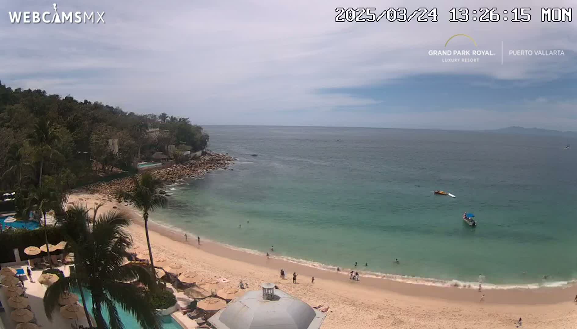 A partly cloudy day shows a tranquil beach scene with clear, teal water, a sandy shoreline, and a resort with swimming pools nestled amongst palm trees.