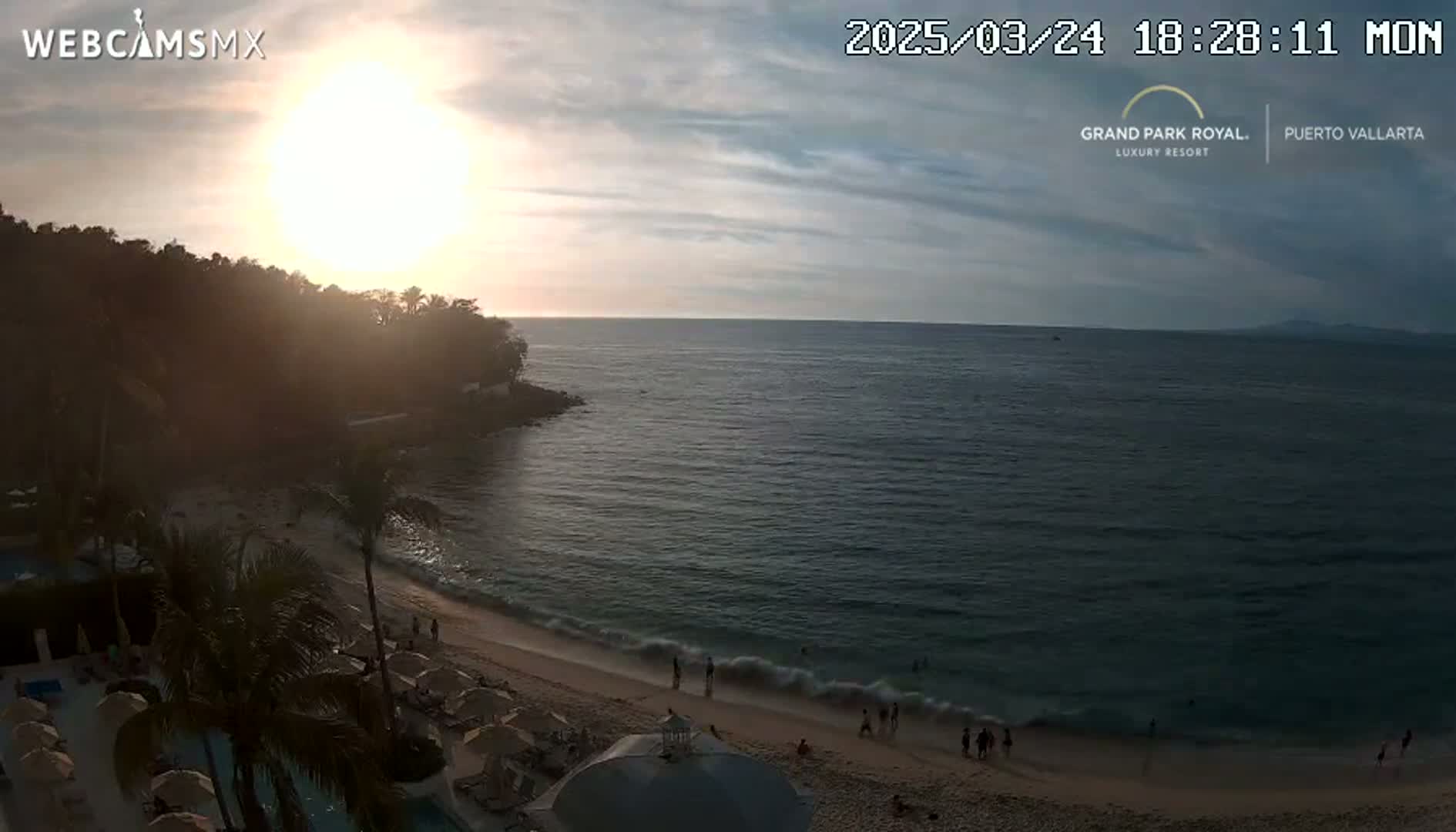 A beach with calm waves at sunset, overlooked by palm trees and a resort.