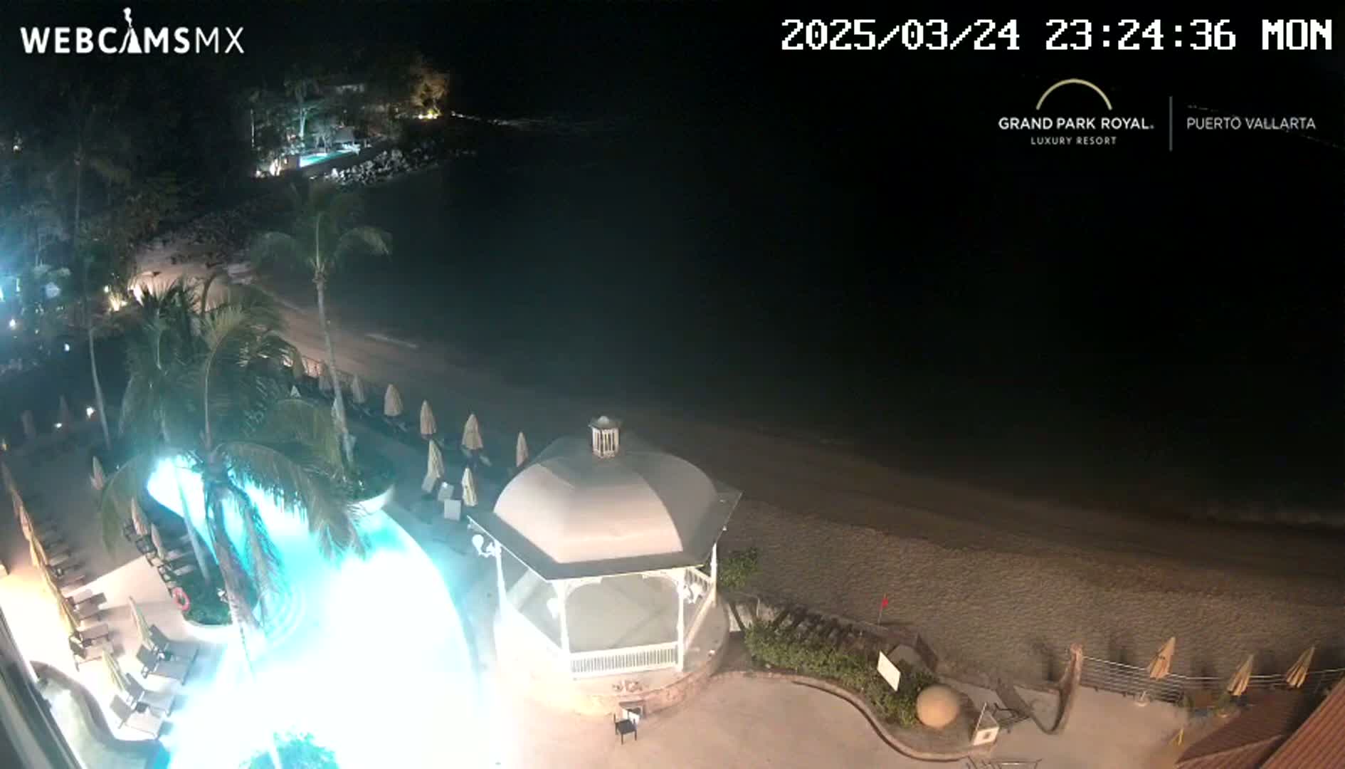 A nighttime overhead view of a resort area shows a pool, beach, and gazebo illuminated under a dark sky.