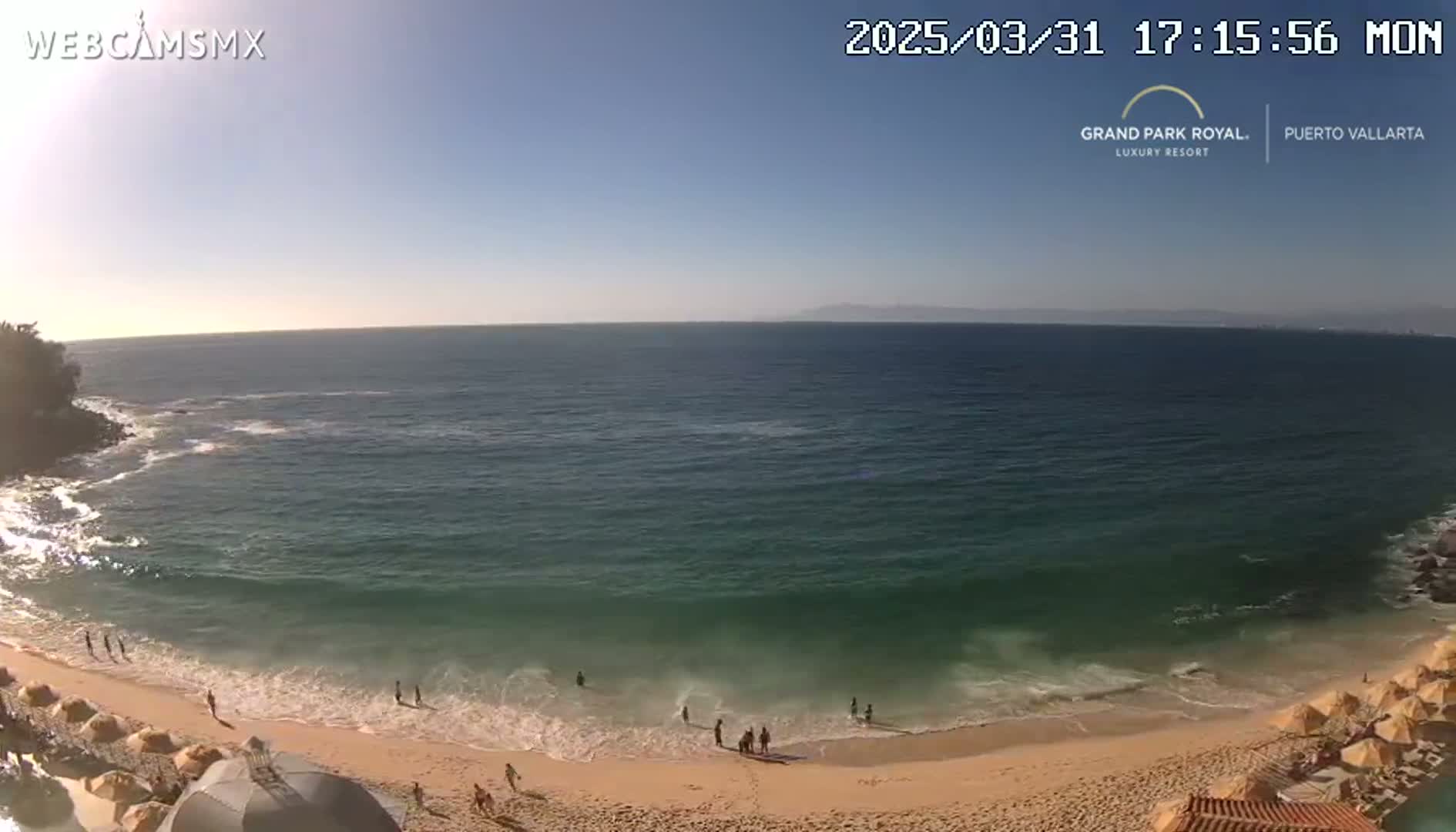 A sandy beach with calm waves and several people swimming in the ocean under a clear, sunny sky.