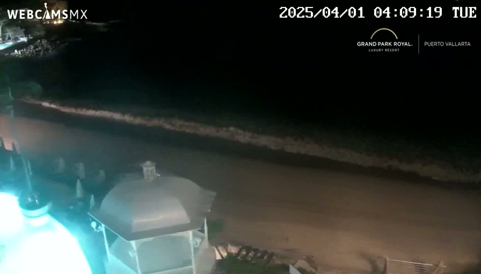 A nighttime view of a sandy beach with gentle waves lapping the shore, seen from an elevated position overlooking a gazebo-like structure.