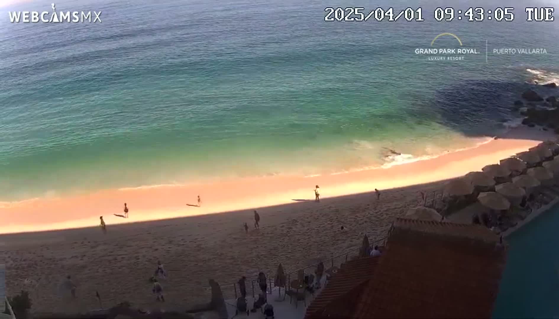 A beach with a few people on it is partially shaded, next to a turquoise ocean on a sunny day.