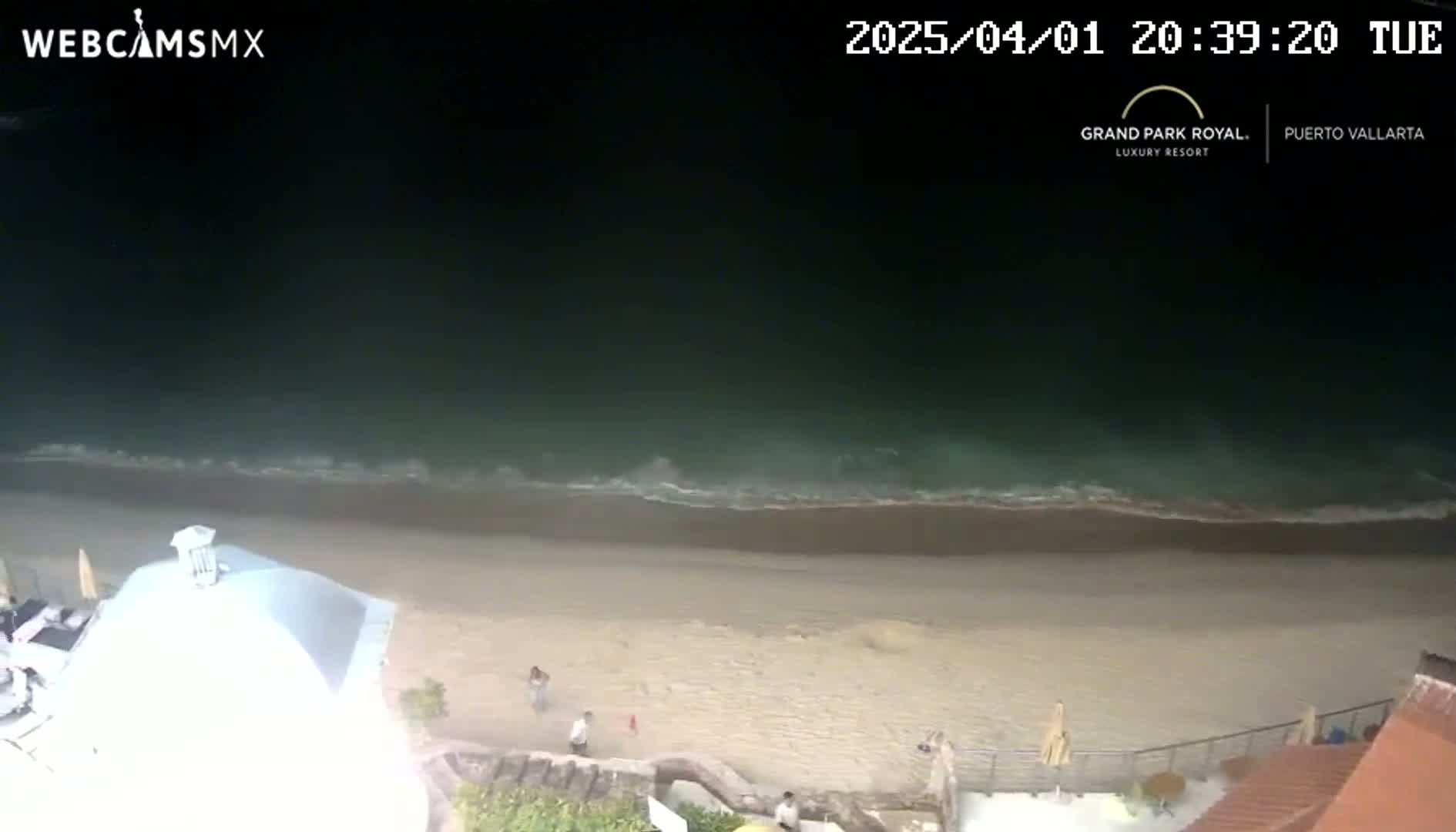A nighttime, high-angle view shows a dark ocean with small waves lapping a sandy beach, with a few people visible on the sand and near a building.