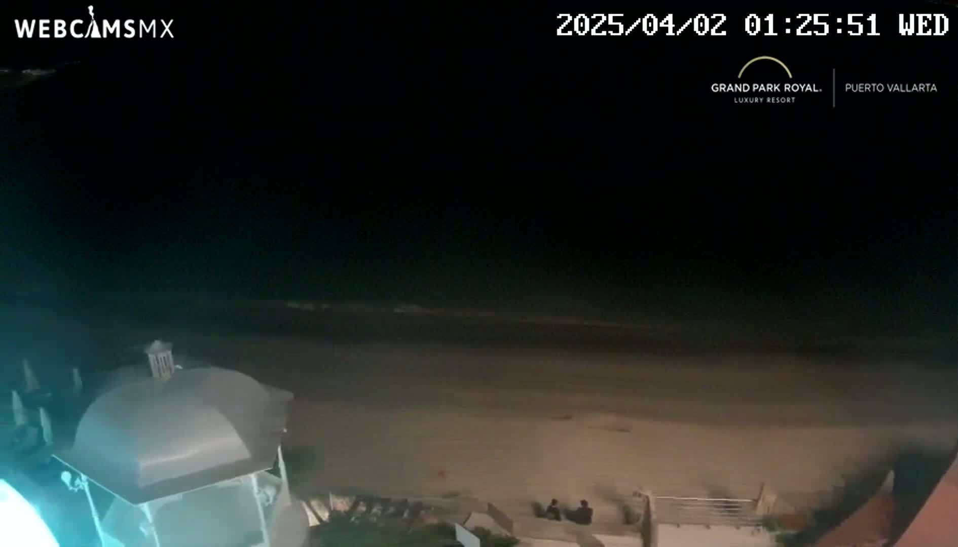 A nighttime view of a dark beach with calm water, a partially visible structure in the foreground, and a few people sitting on the sand.