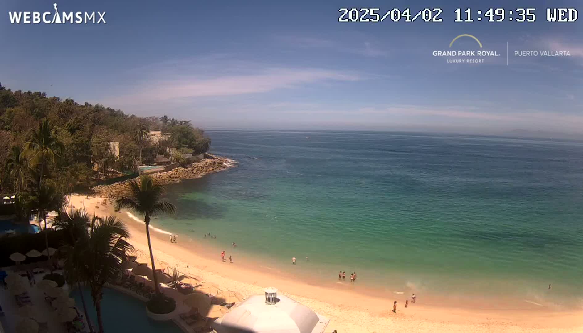A sandy beach with calm, clear turquoise water is nestled in a cove bordered by lush green vegetation and buildings under a mostly sunny sky.