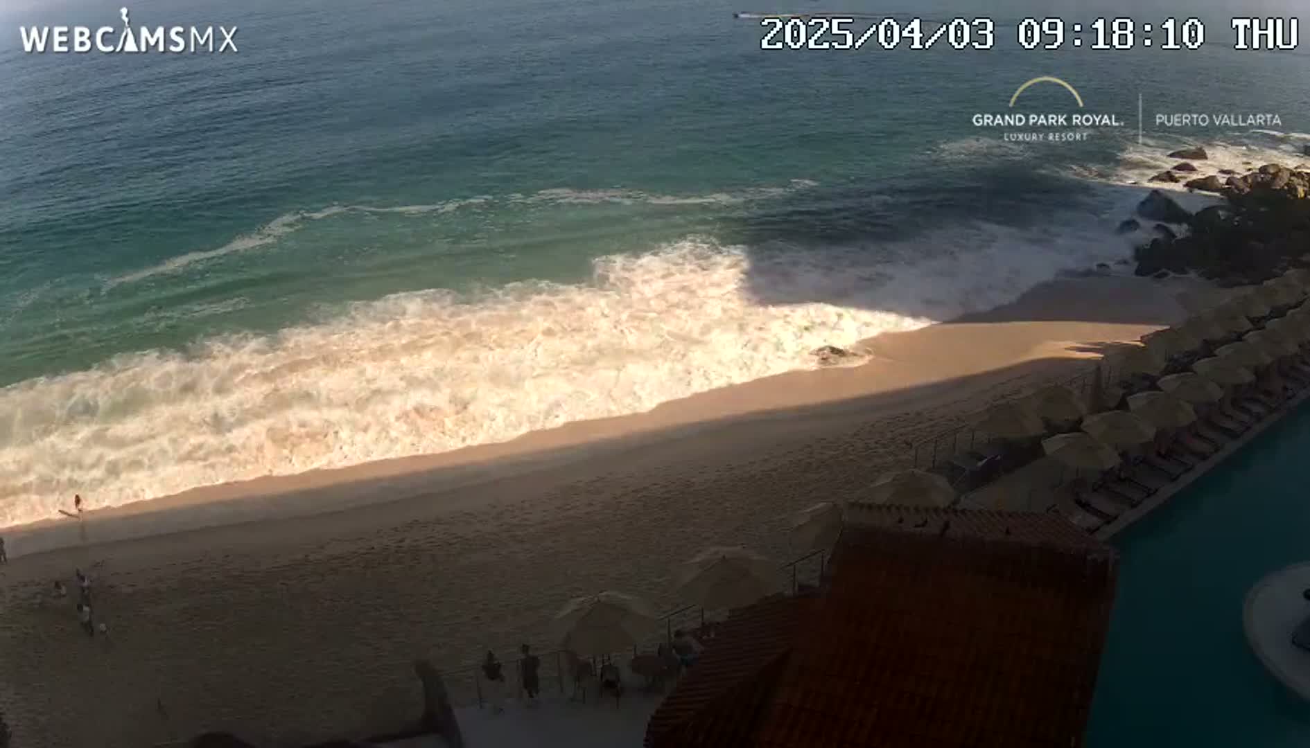 A sandy beach with moderate waves and several beach umbrellas and lounge chairs next to a swimming pool is seen from an elevated vantage point on a sunny day.