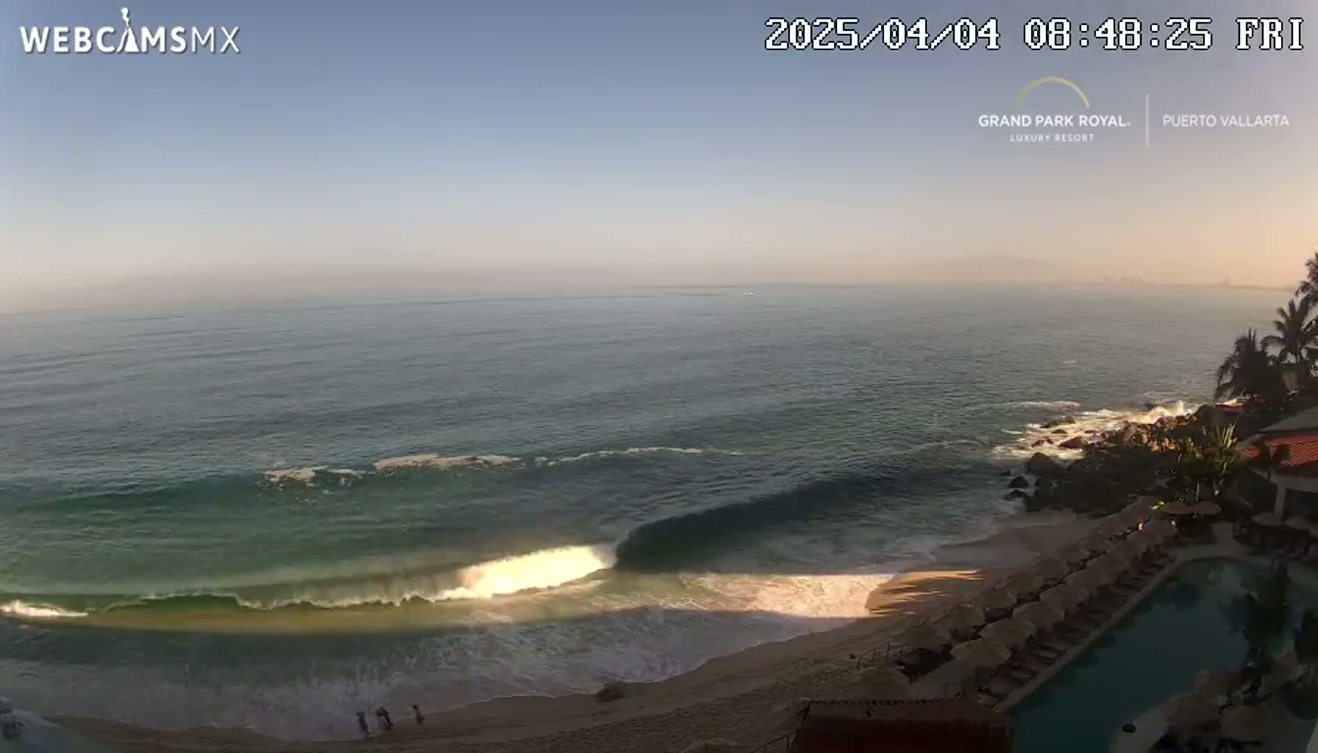 An ocean beach with waves rolling onto a sandy shore next to a resort with a swimming pool under a clear, bright sky.