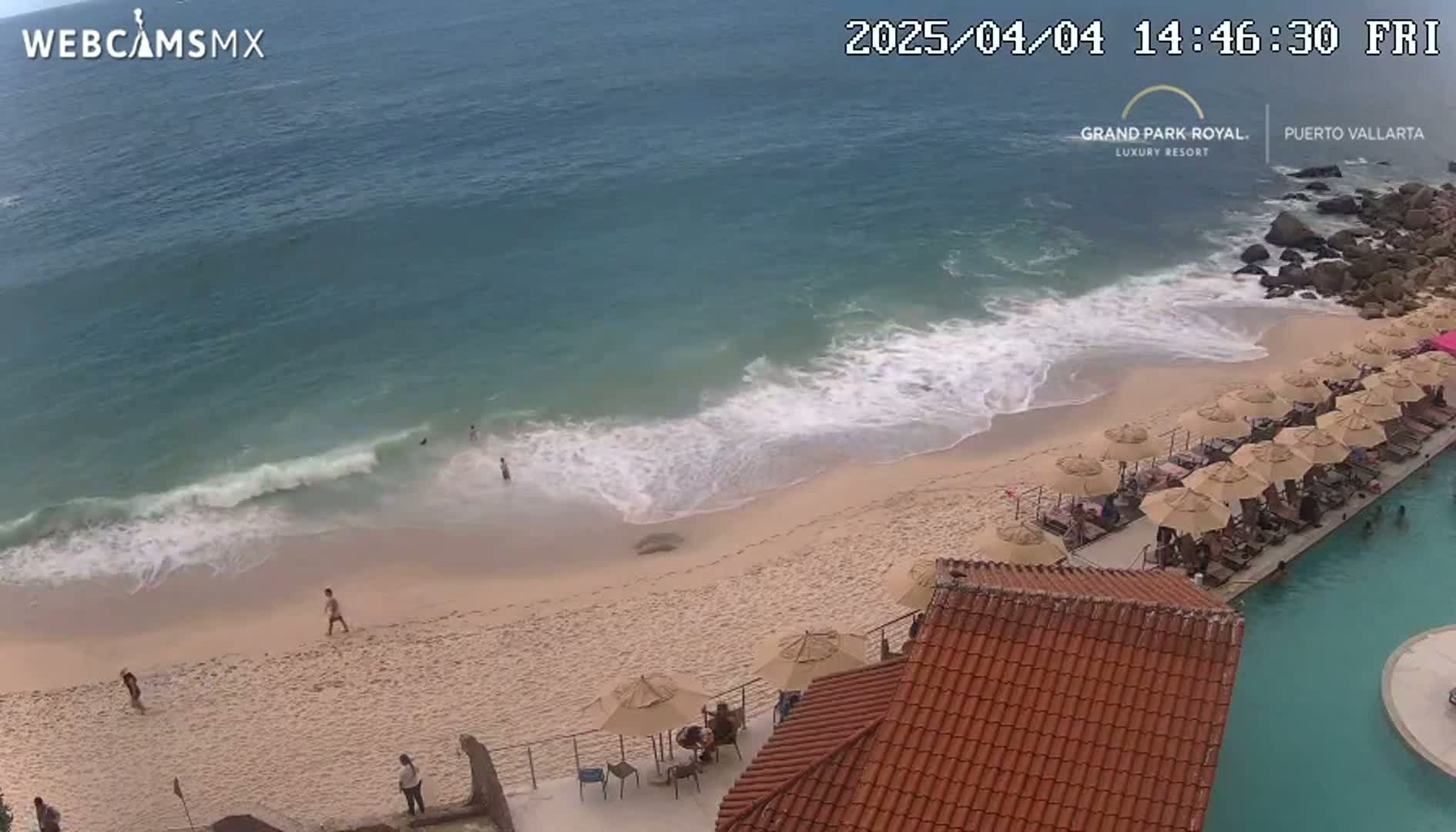 An aerial view shows a sandy beach with a few people on it, gentle waves lapping the shore, numerous umbrellas lining a pool and patio area next to the beach, all under an overcast sky.
