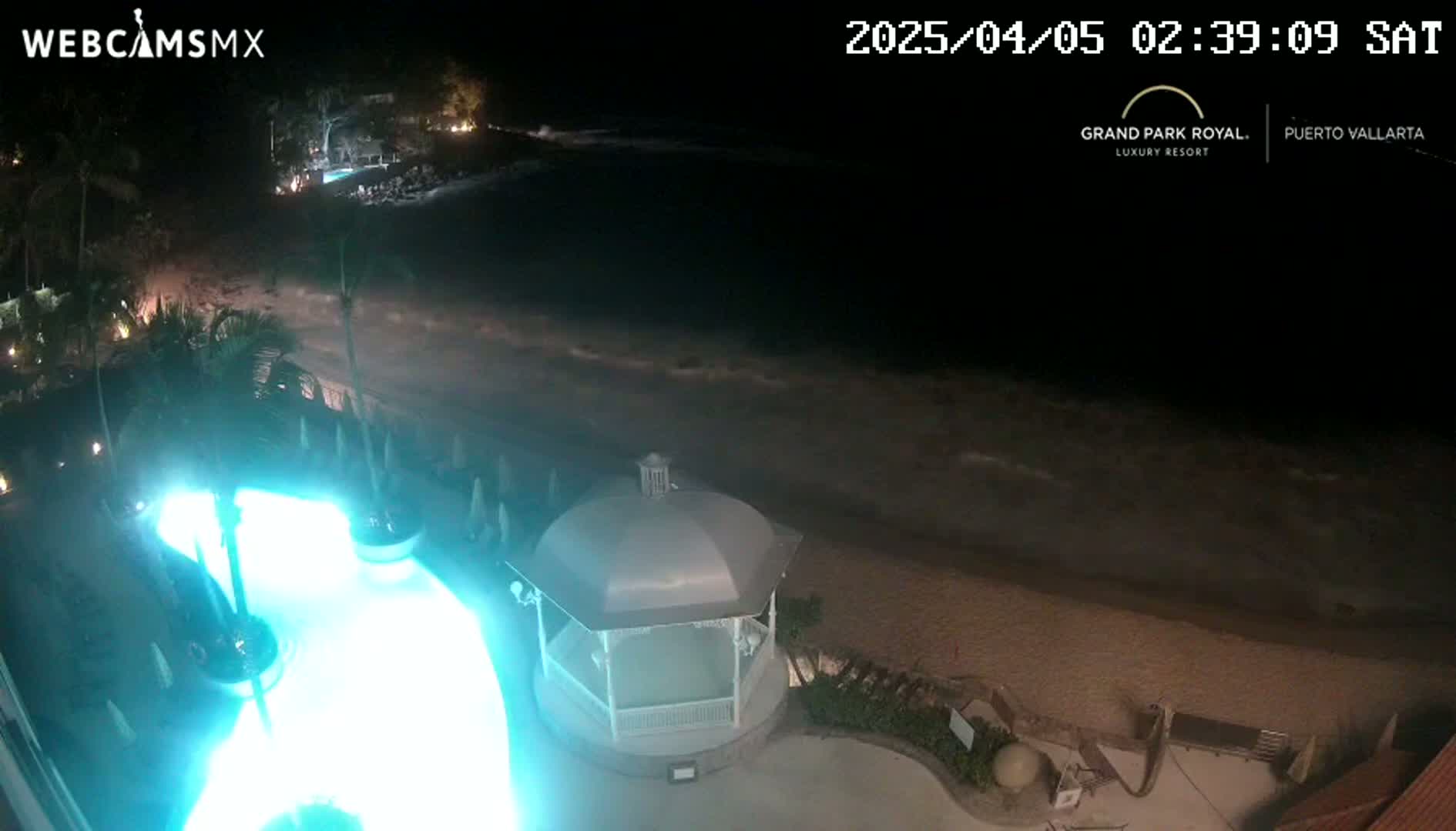 A nighttime view of a resort pool illuminated in turquoise, a beach with gentle waves, and a gazebo overlooking the ocean.