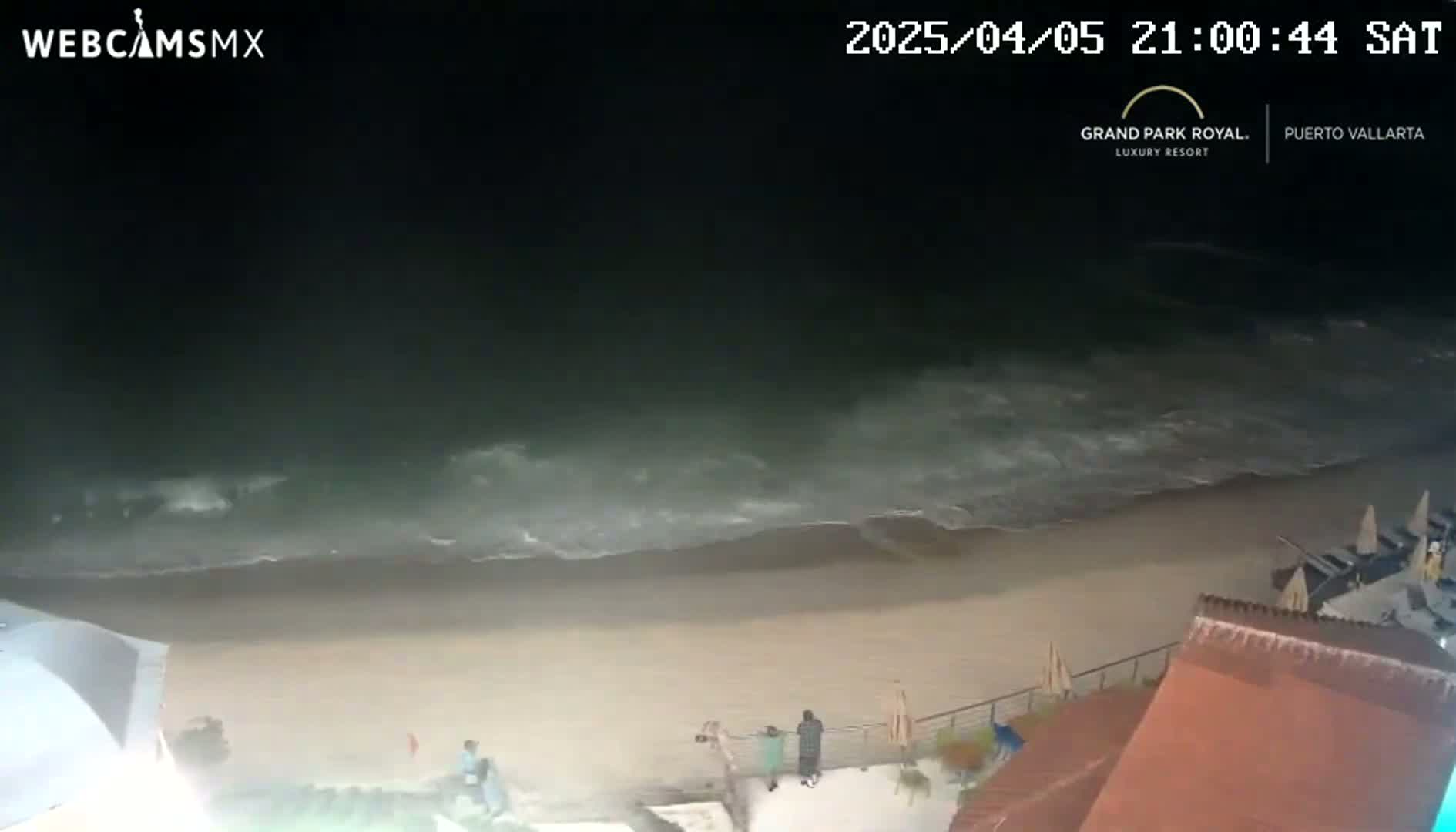 At night, moderately sized waves crash on a sandy beach near a resort, under a dark sky.