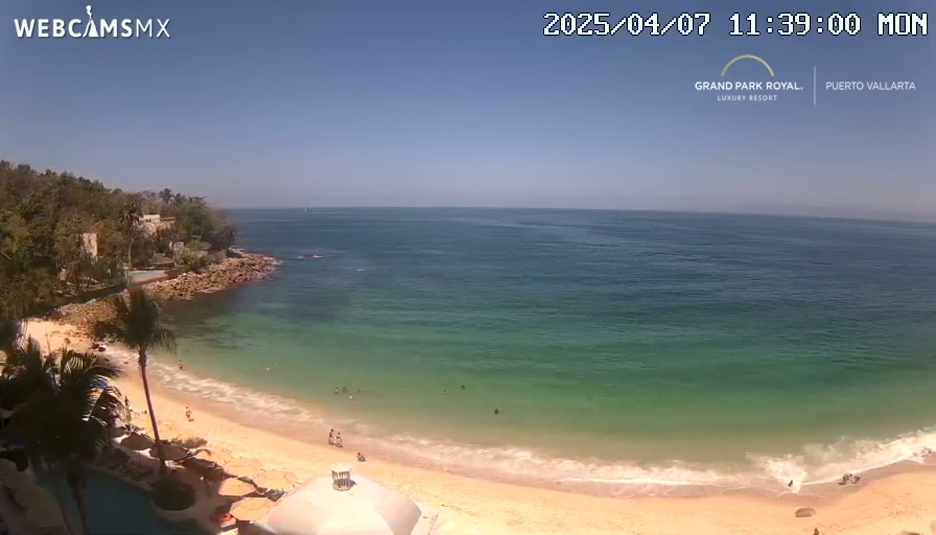 A sandy beach with calm, clear turquoise water laps gently at the shore under a bright sunny sky,  backed by tropical vegetation and rocky cliffs.