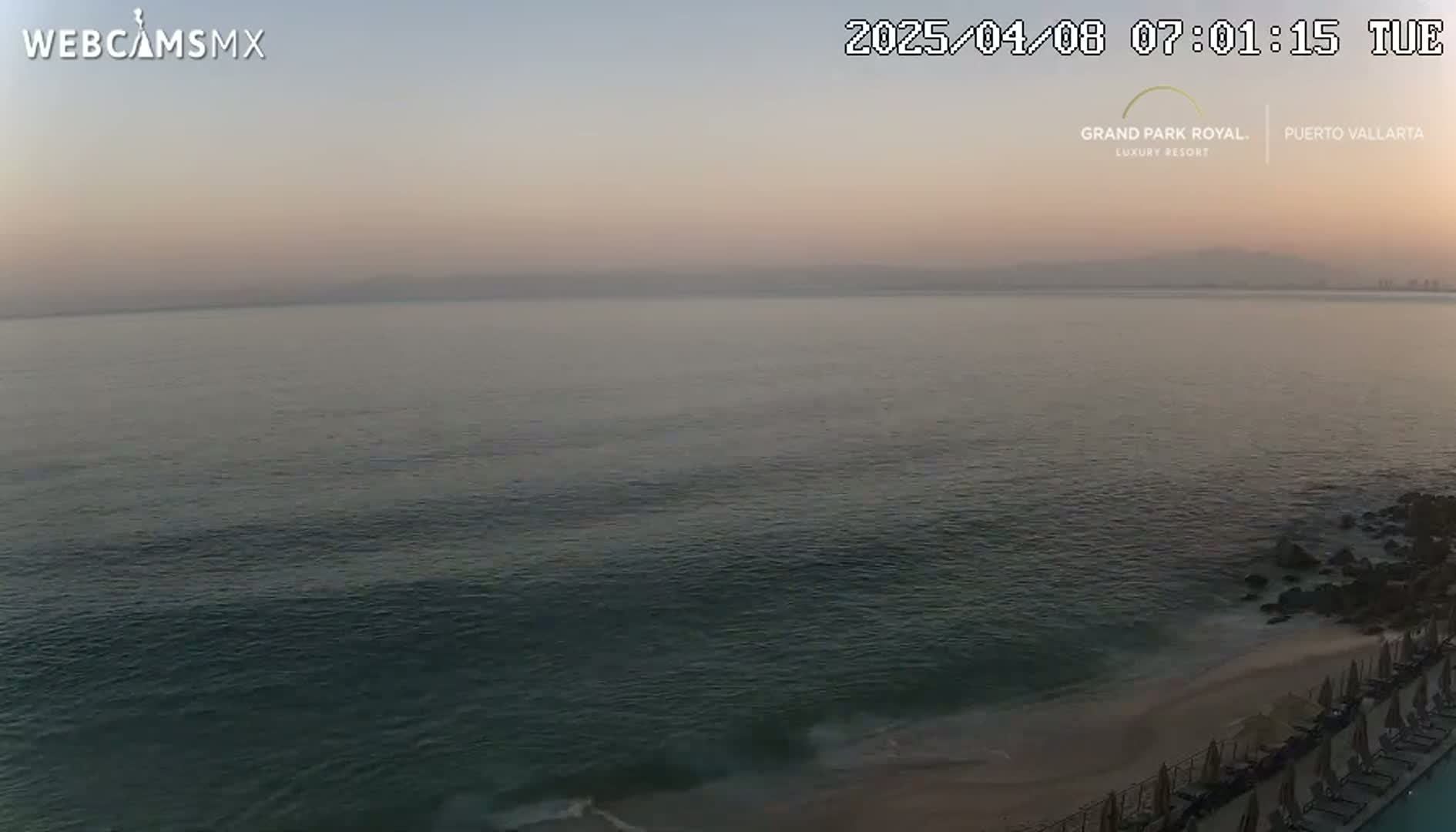A hazy sunrise over a calm ocean, with a sandy beach and resort chairs visible in the foreground.