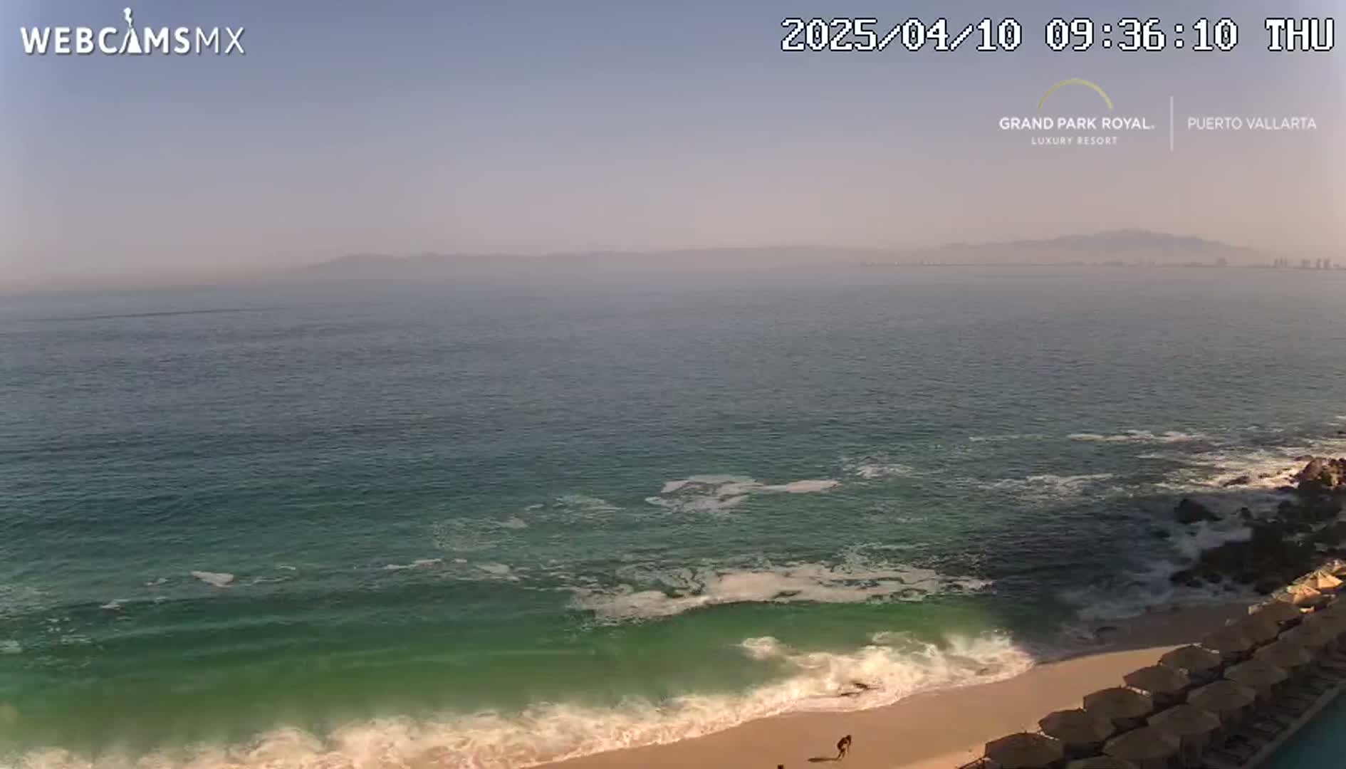 A hazy, sunny day overlooking a beach with gentle waves lapping the shore, and a row of beach umbrellas visible in the foreground.