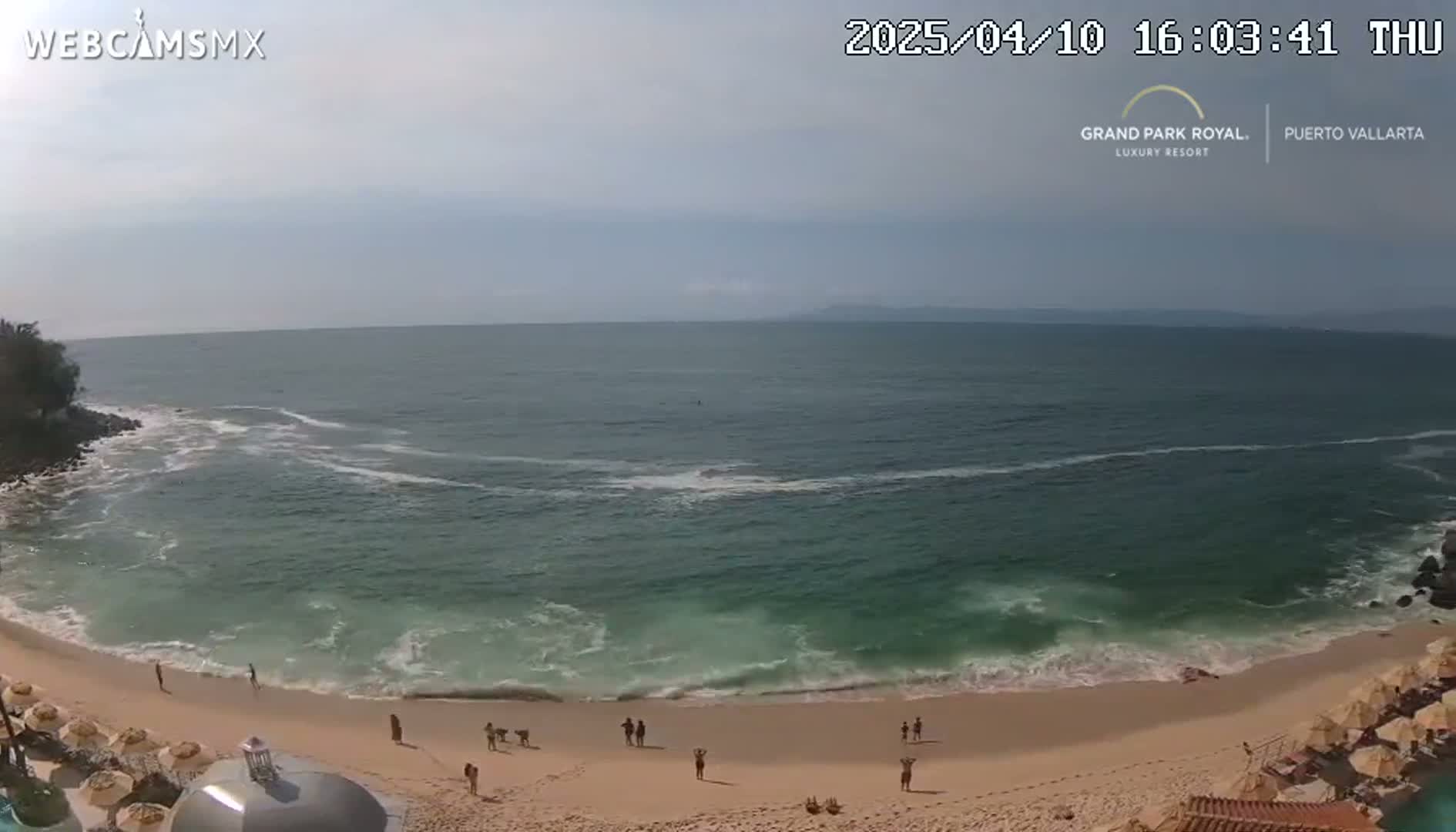 A mostly sunny day shows a beach with a few people, light waves lapping a sandy shore, and a calm ocean extending to a hazy horizon.